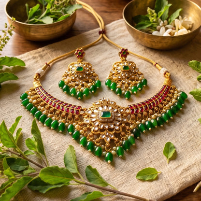 An ornate gold-toned traditional kundan necklace and matching earrings set with green beads, white stones, and red gemstone accents, arranged on a beige fabric surface with decorative green leaves and brass bowls in the background.