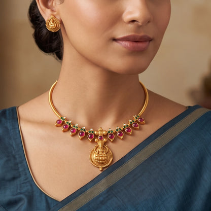 model wearing matte antique gold finish Hasli choker necklace featuring alternating pink and green cabochon stones and a central Goddess Lakshmi pendant with matching earrings.