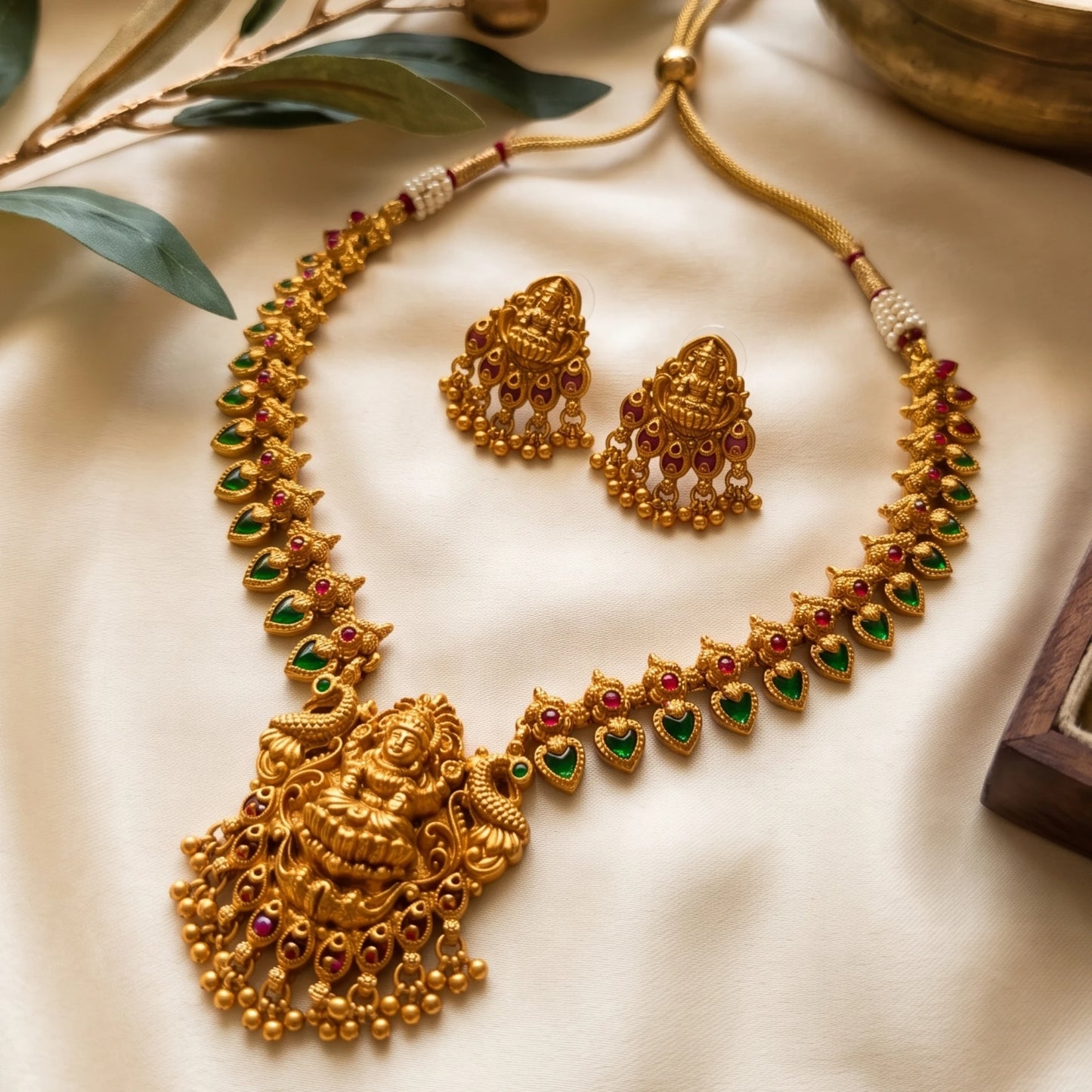 Gold-toned traditional jewelry set featuring a necklace with a large deity pendant, matching earrings, and heart-shaped motifs accented with green and red stones, arranged on cream fabric with decorative leaves.