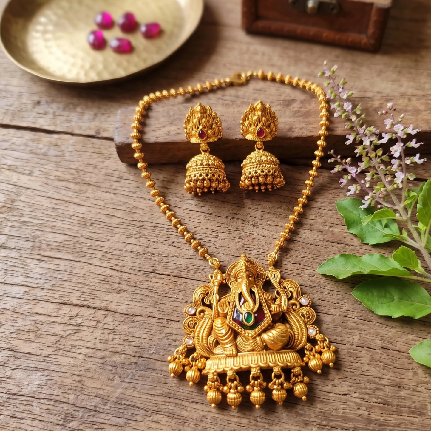 Traditional Indian matte antique gold temple necklace featuring a Lord Ganesha pendant and matching Jhumka earrings on a rustic wooden table next to a fresh Tulsi stem with small flowers.