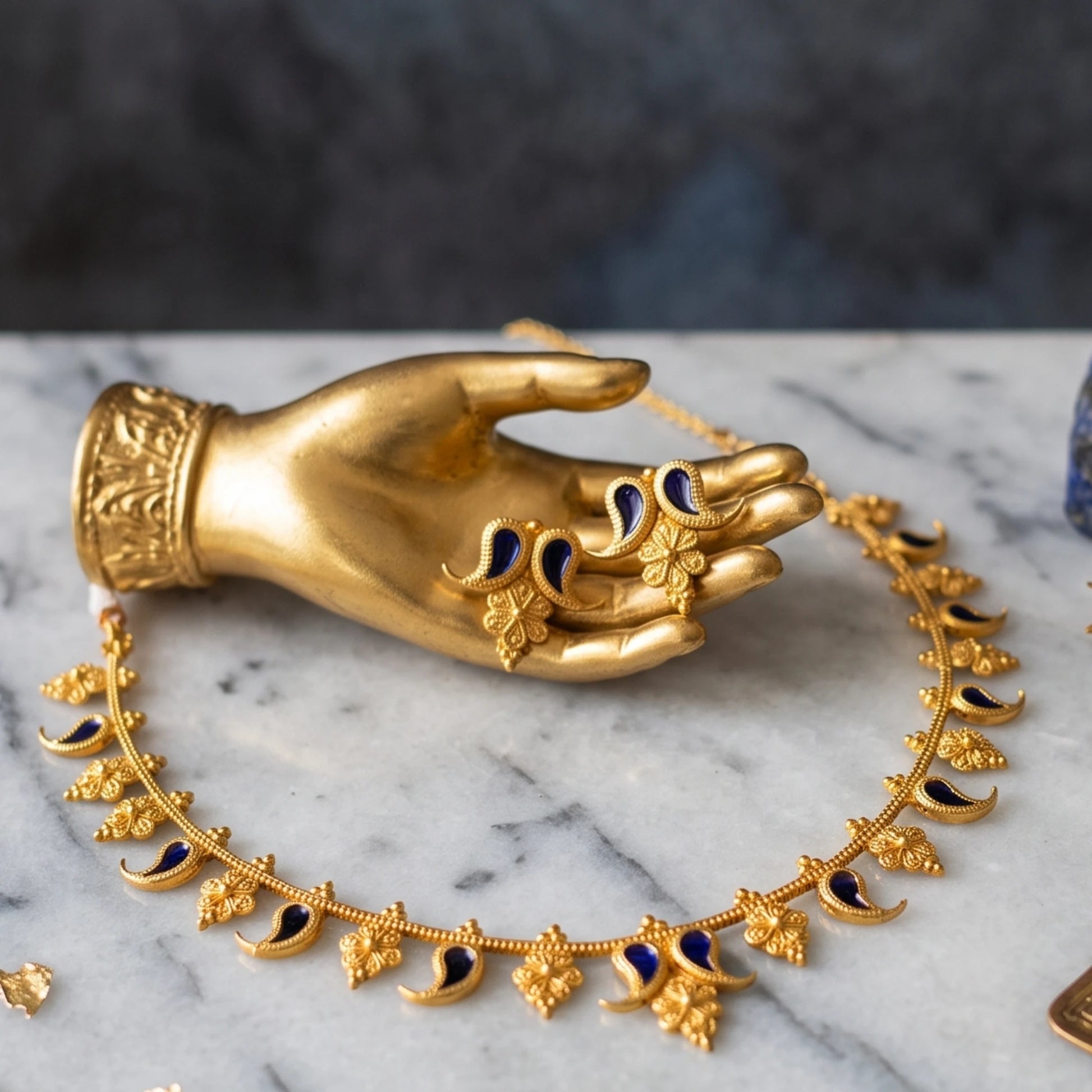 Gold-toned ornate jewelry displayed on a white marble surface with a metallic hand-shaped stand. The set includes a necklace with leaf and crescent motifs and a earring with floral and paisley details, both accented with dark blue enamel.