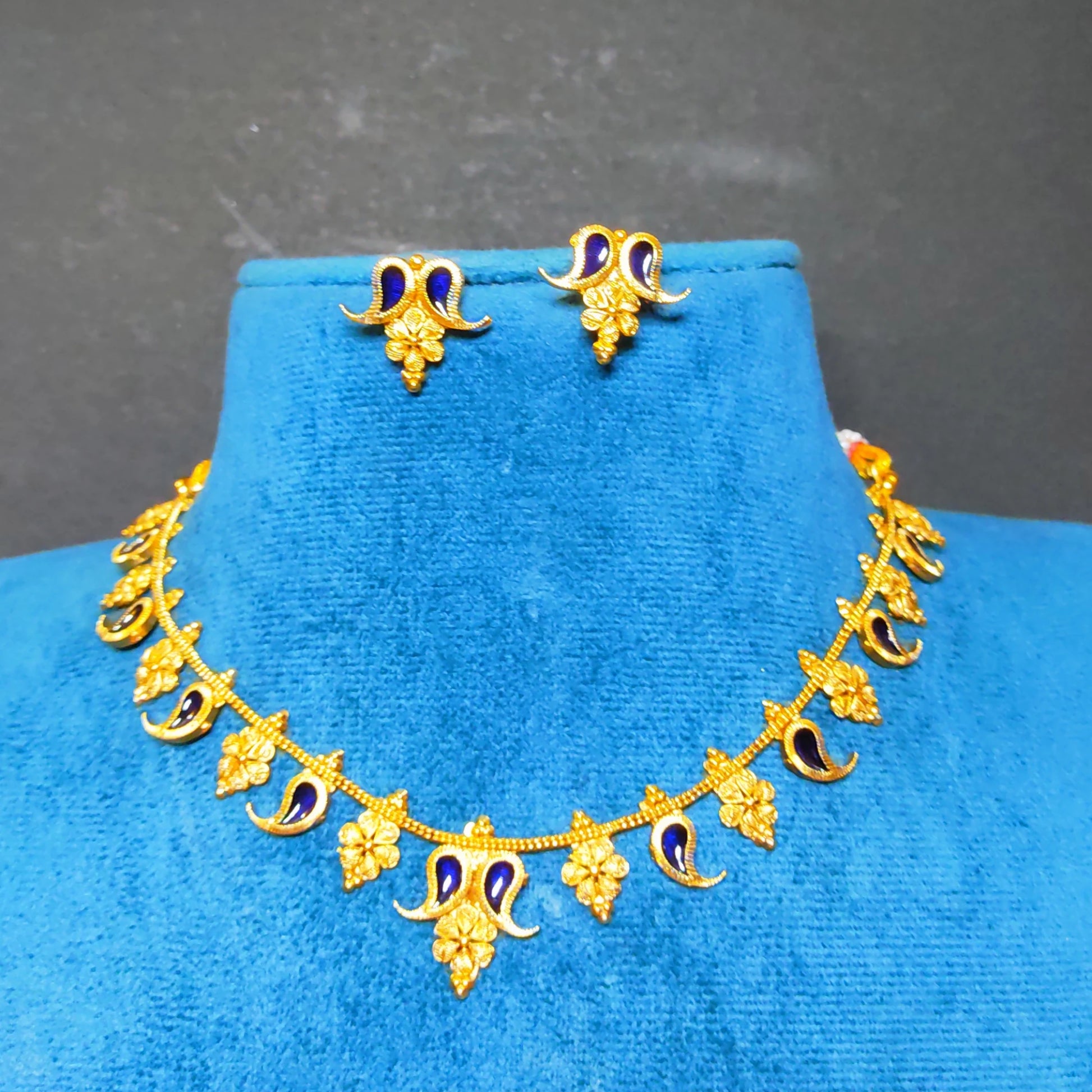 Gold-toned jewelry set including a necklace and matching earrings with blue enamel accents, displayed on a bright blue velvet jewelry bust against a dark gray background.