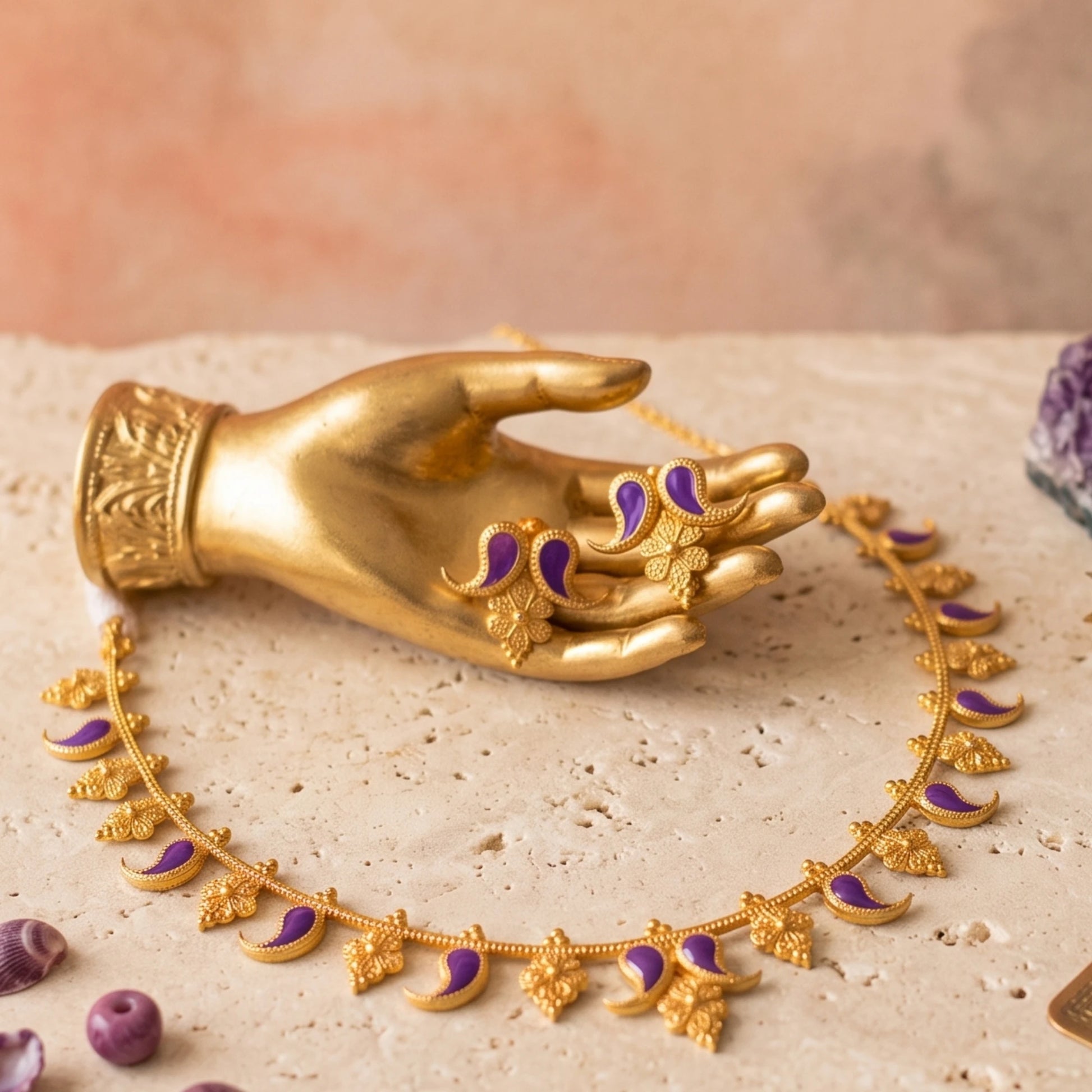 Gold-toned jewelry set displayed on a beige stone surface, including a necklace with crescent and floral motifs and matching earring, both accented with purple enamel, shown with a gold toned mannequin hand.