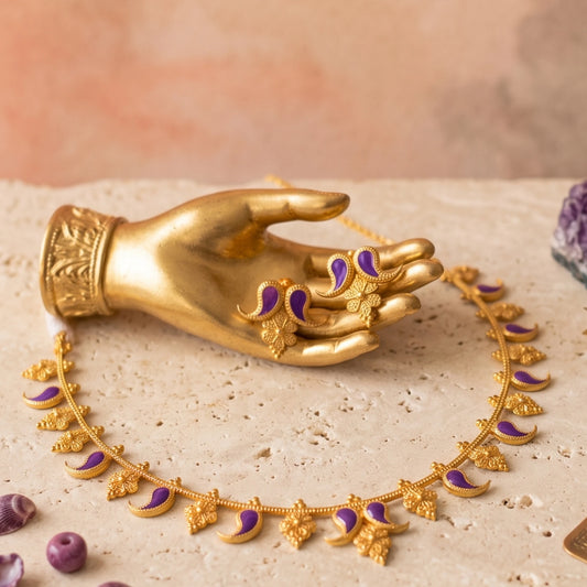 Gold-toned jewelry set displayed on a beige stone surface, including a necklace with crescent and floral motifs and matching earring, both accented with purple enamel, shown with a gold toned mannequin hand.