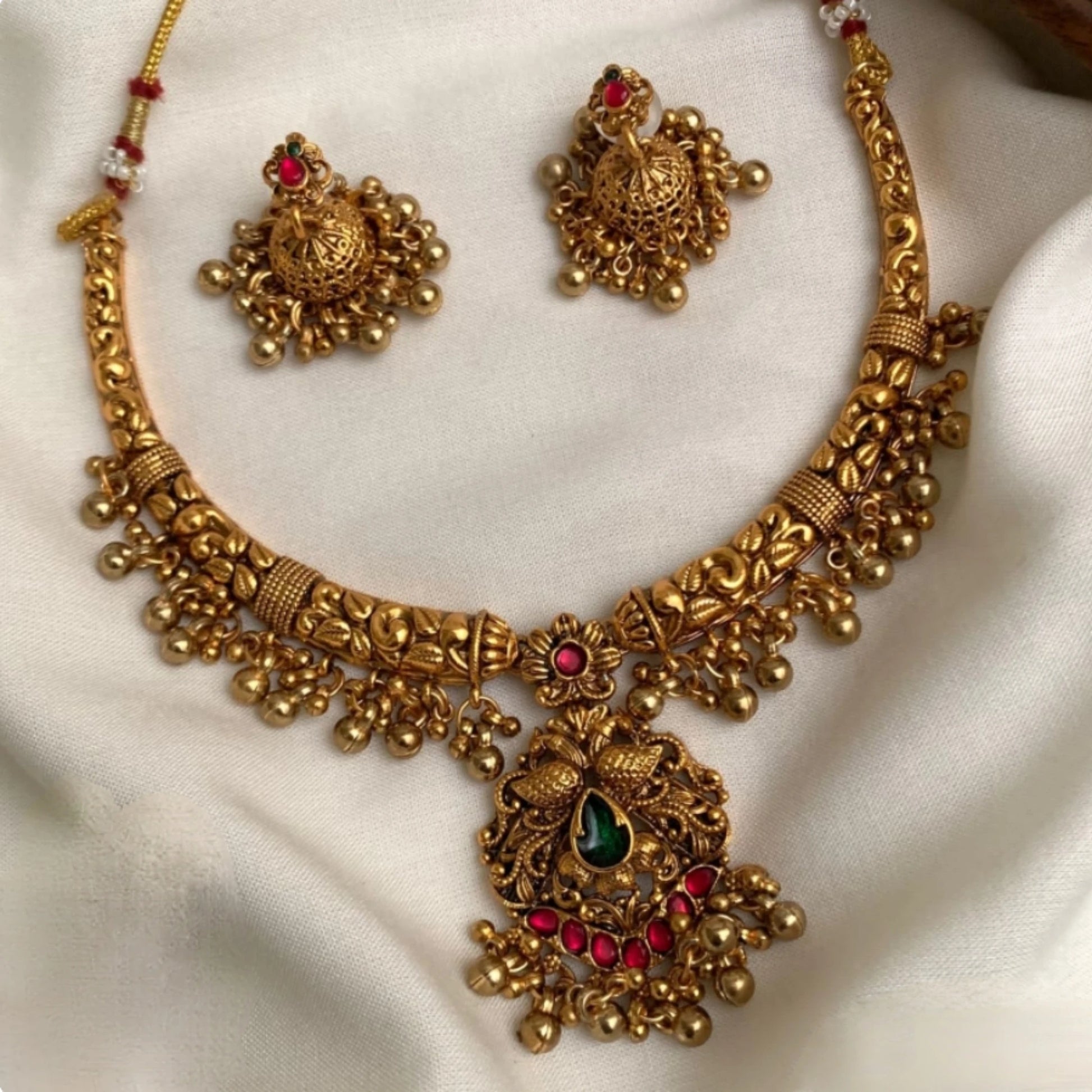The set includes an antique matte gold finish choker necklace and matching jhumkas featuring detailed peacock carvings, ruby red and emerald green gemstones, and hanging gold toned clusters.