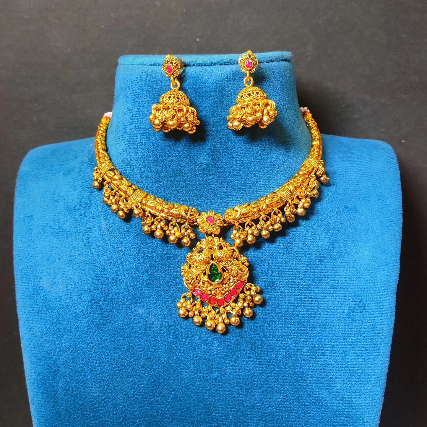 The set includes an antique matte gold finish choker necklace and matching jhumkas featuring detailed peacock carvings, ruby red and emerald green gemstones, and hanging gold toned clusters