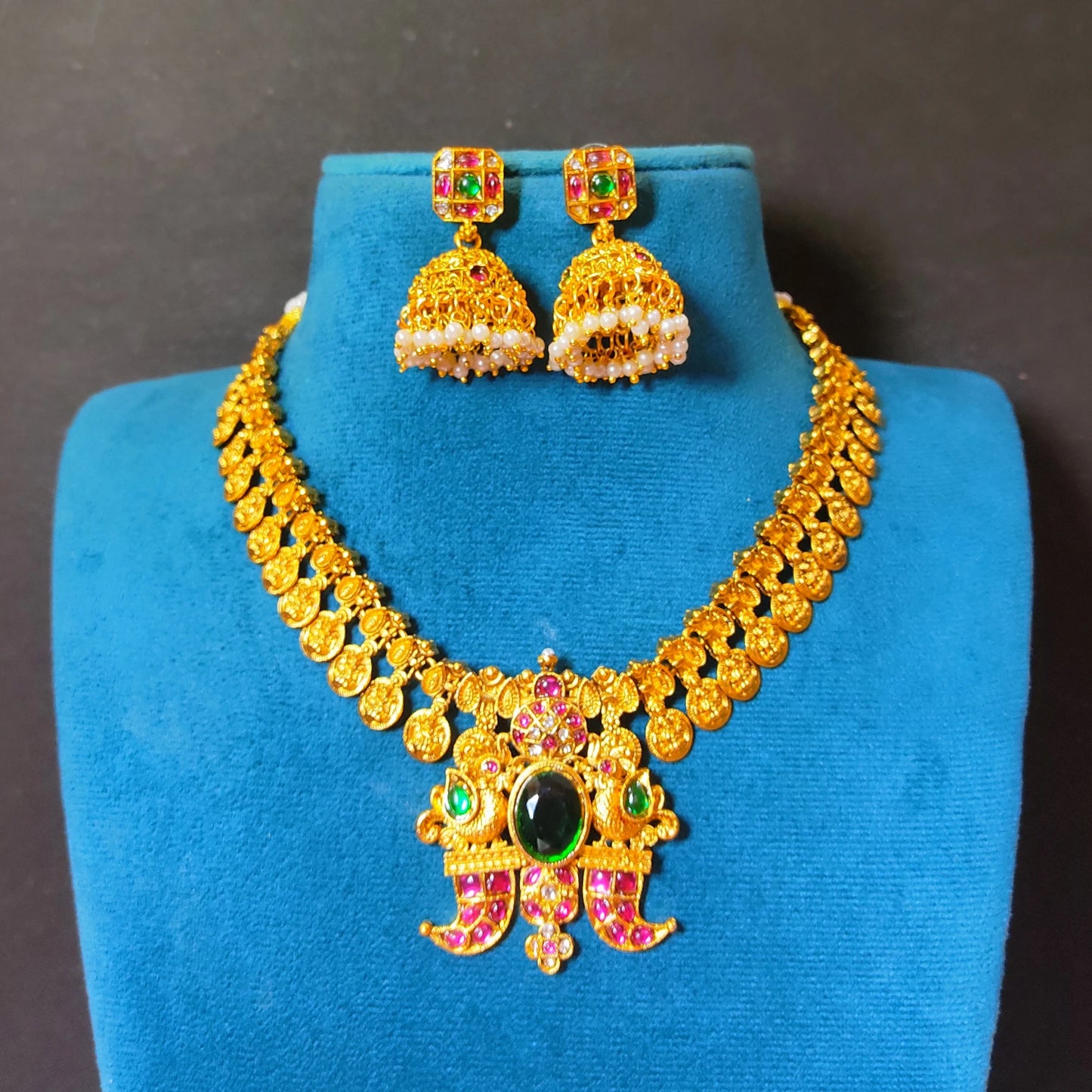 Close-up of traditional South Indian matte gold toned temple jewelry set. The necklace features Laxmi coin chain and a detailed twin-peacock pendant with a large oval green emerald and pink rubies and comes with matching jimikkis/jhumkas