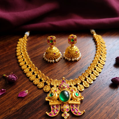 Close-up of traditional South Indian matte gold toned temple jewelry set. The necklace features Laxmi coin chain and a detailed twin-peacock pendant with a large oval green emerald and pink rubies and comes with matching jimikkis/jhumkas