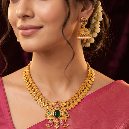 Close-up of a model wearing traditional South Indian matte gold toned temple jewelry set. The necklace features Laxmi coin chain and a detailed twin-peacock pendant with a large oval green emerald and pink rubies and comes with matching jimikkis/jhumkas