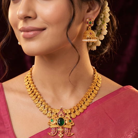 Close-up of a model wearing traditional South Indian matte gold toned temple jewelry set. The necklace features Laxmi coin chain and a detailed twin-peacock pendant with a large oval green emerald and pink rubies and comes with matching jimikkis/jhumkas
