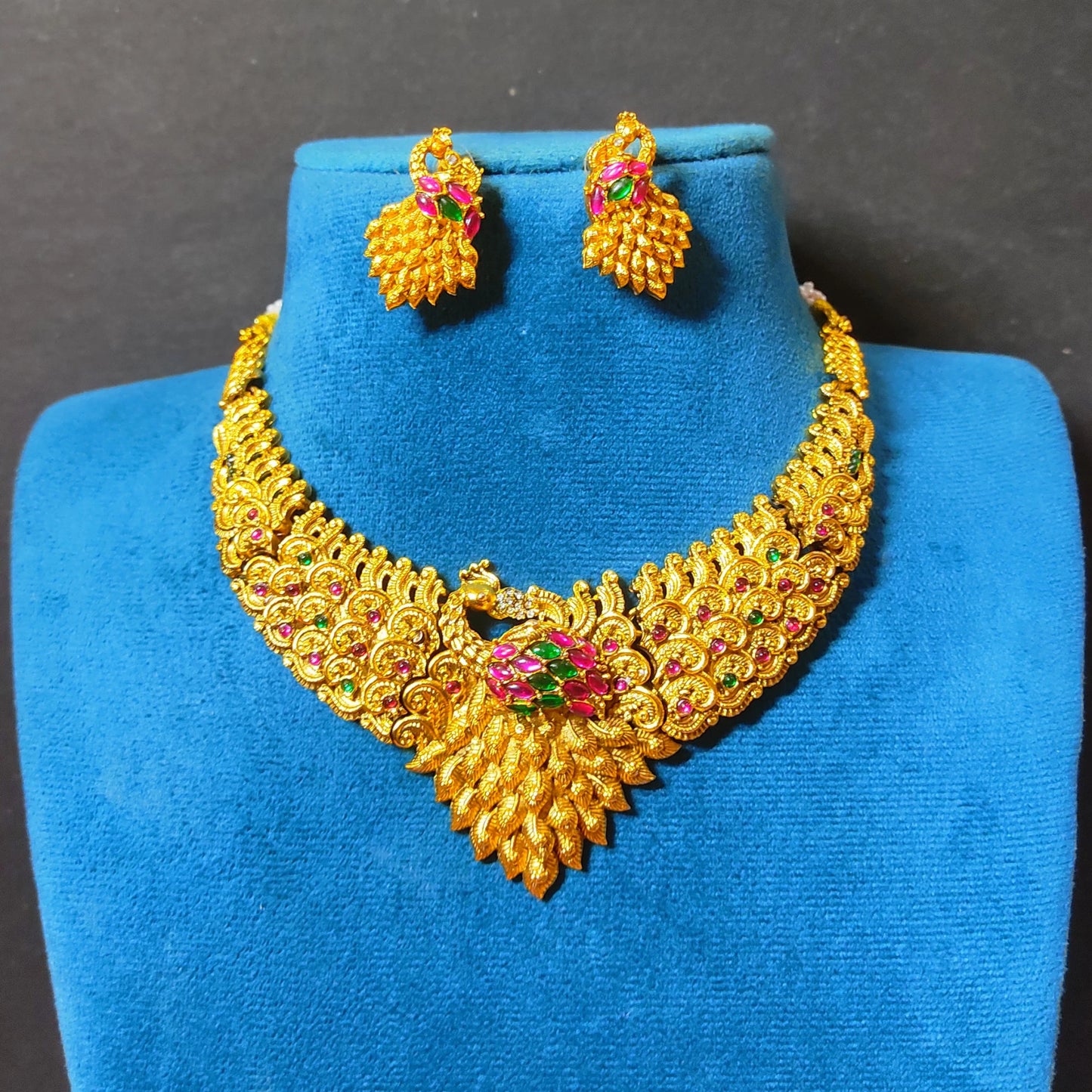 Traditional Indian matte antique gold peacock choker necklace and matching statement stud earrings beautifully arranged on a soft, blue mannequin