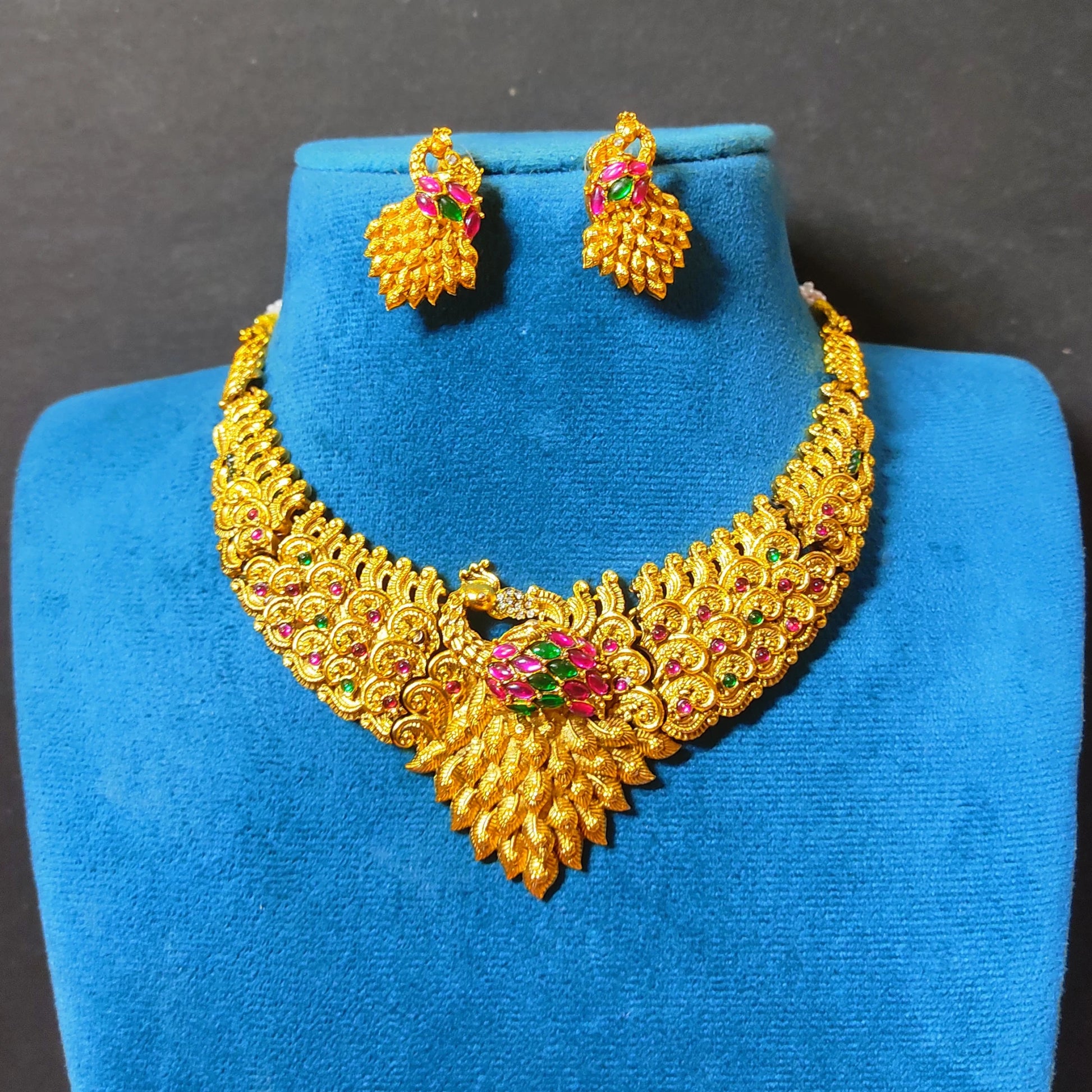 Traditional Indian matte antique gold peacock choker necklace and matching statement stud earrings beautifully arranged on a soft, blue mannequin