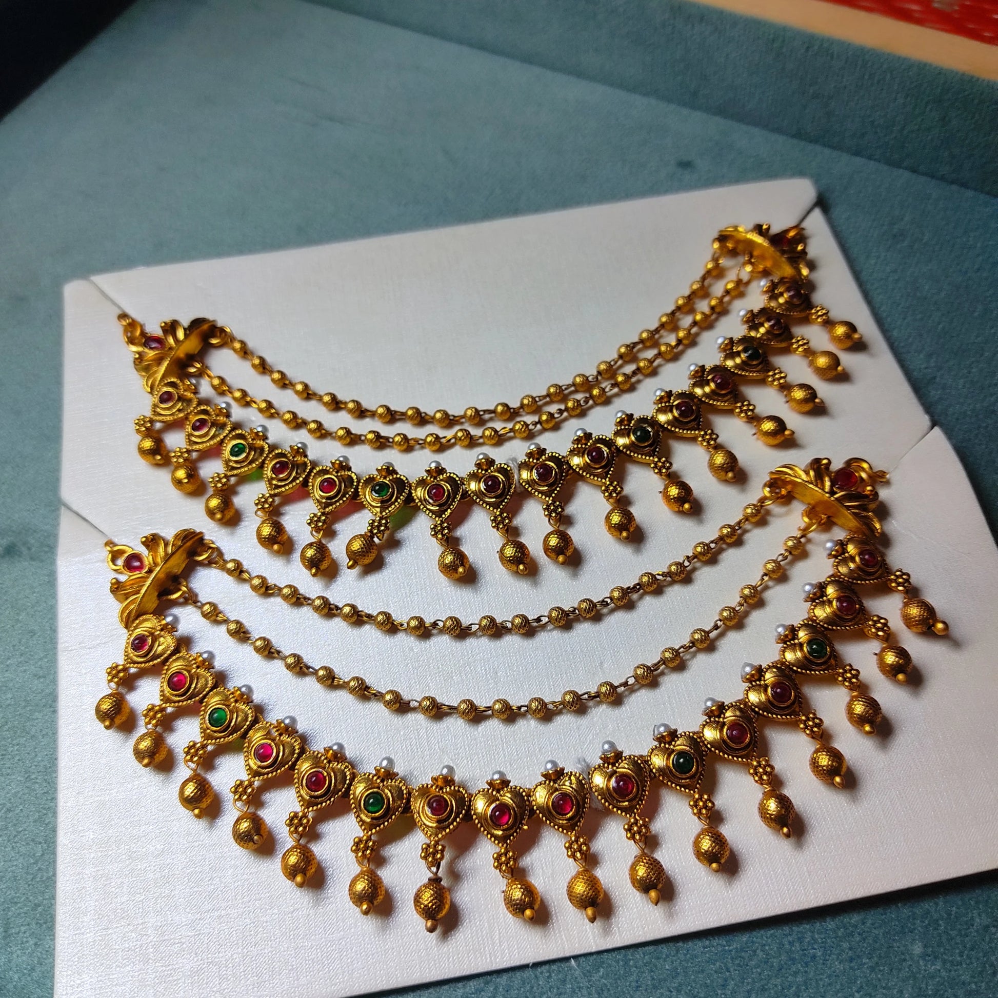 A pair of traditional Indian 3-layer antique gold mattal ear chains with ruby red and emerald green kemp stones, displayed beautifully