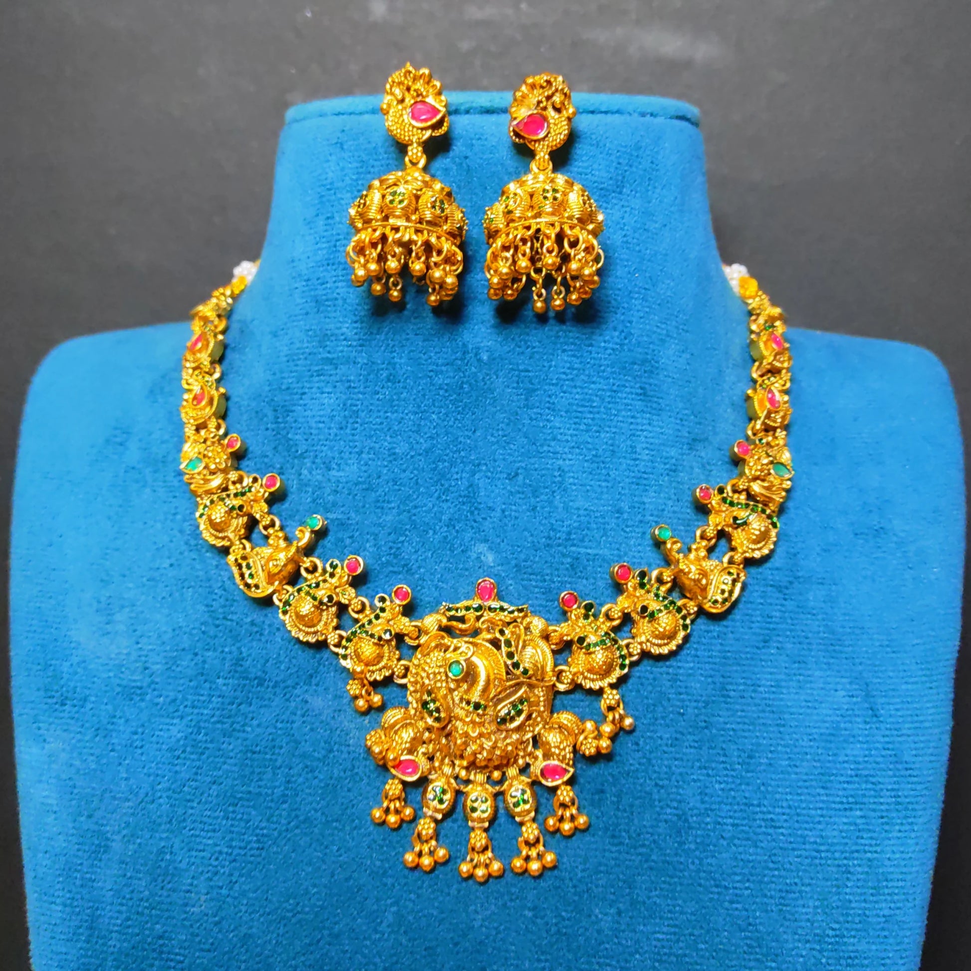 Antique Gold-toned traditional jewelry set featuring a necklace and matching earrings displayed on a blue velvet jewelry bust. The pieces are decorated with red and green stone accents and has peacock motif as the central pendant