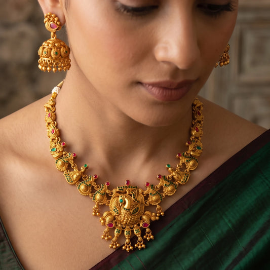 A close-up of a woman wearing an ornate antique gold-toned traditional necklace with matching dangling earrings. The jewelry is decorated with small red and green stone accents and has peacock/mayil as the central pendant.