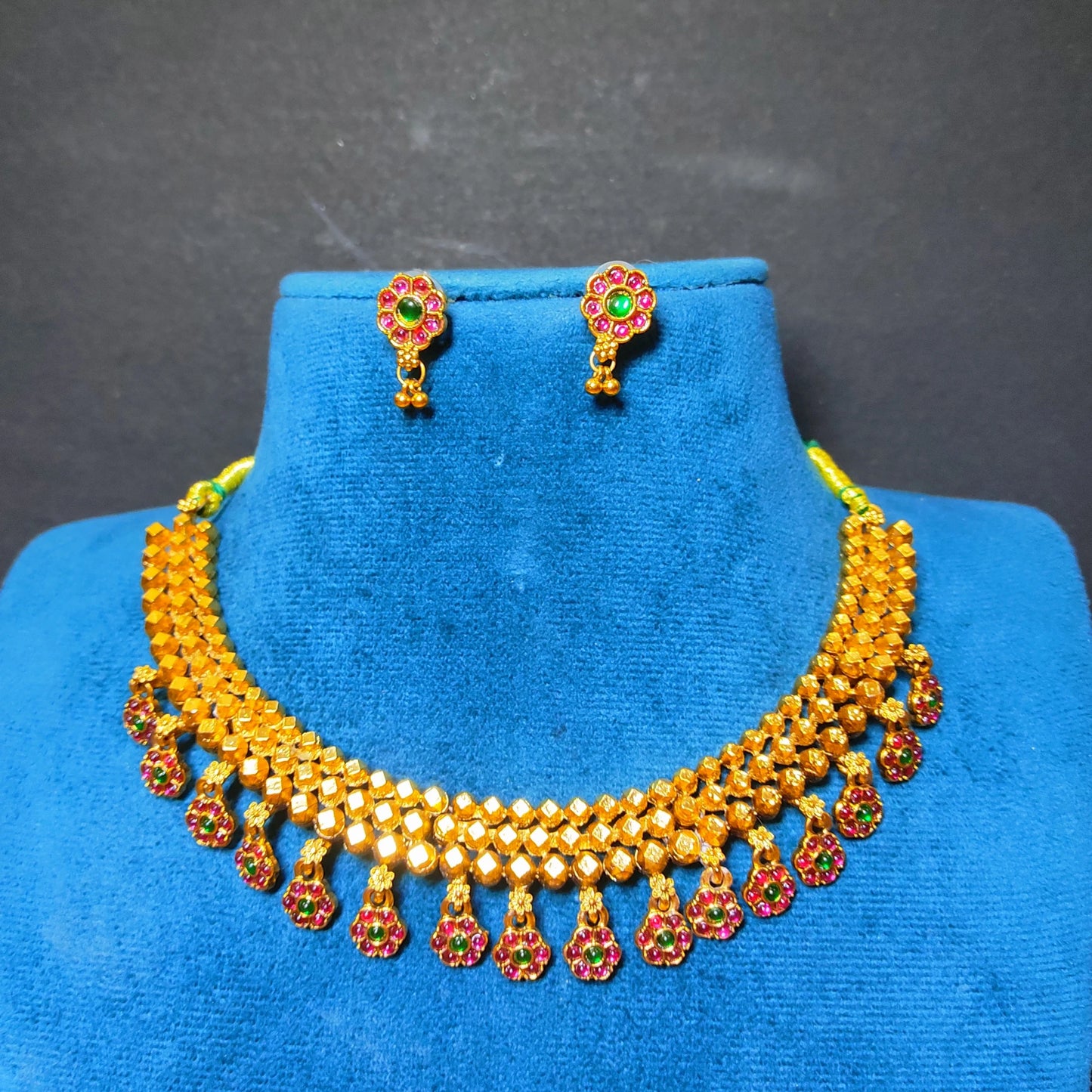 Gold-tone traditional necklace and matching stud earrings with red and green stone accents, displayed on a blue velvet jewelry bust against a dark gray background.