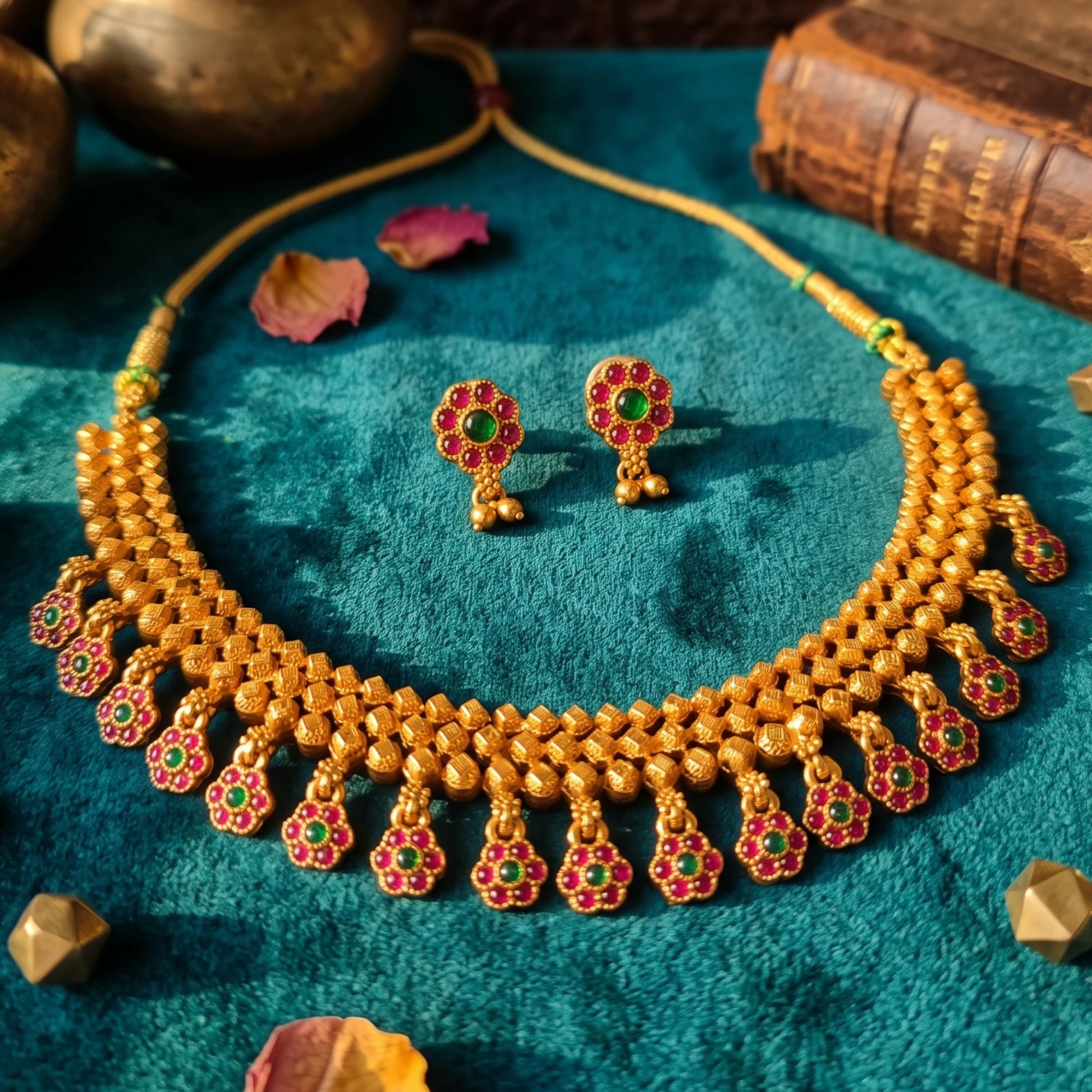 A gold-toned traditional necklace and matching earrings with red and green stone accents, arranged on a teal textured fabric with decorative props.