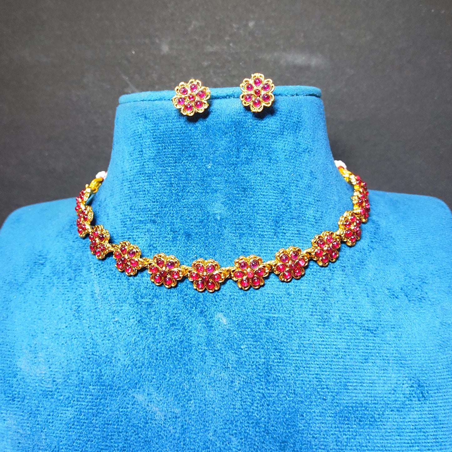Antique Gold finish jewelry set with red gemstone floral motifs, including a necklace and matching stud earrings, displayed on a blue velvet jewelry bust against a dark gray background.