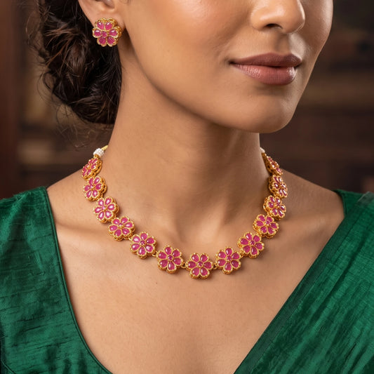 A close-up of a woman wearing a antique gold finish gold and pink floral necklace with matching flower-shaped earrings, paired with a green top.