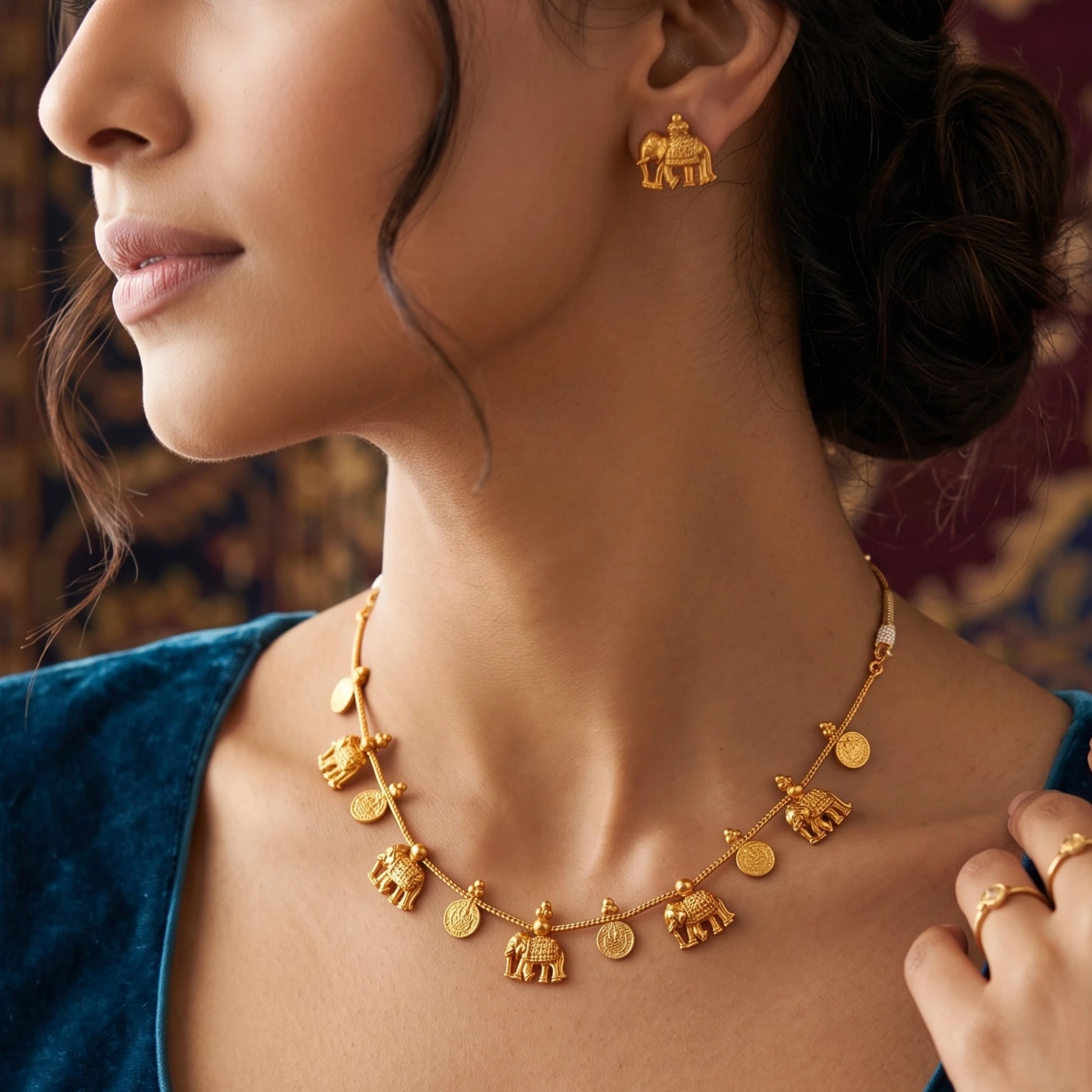 A close-up lifestyle shot of a model wearing a gold toned necklace and matching gold toned earring, both featuring elephant and coin charms, set against a warm, blurred background.