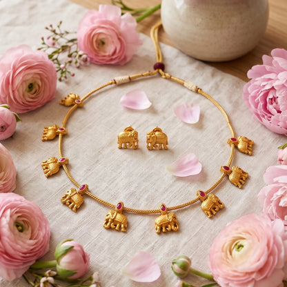 Gold toned elephant jewelry set with a necklace and matching earrings, decorated with small pink gemstone accents, arranged on a beige fabric with pink flowers.