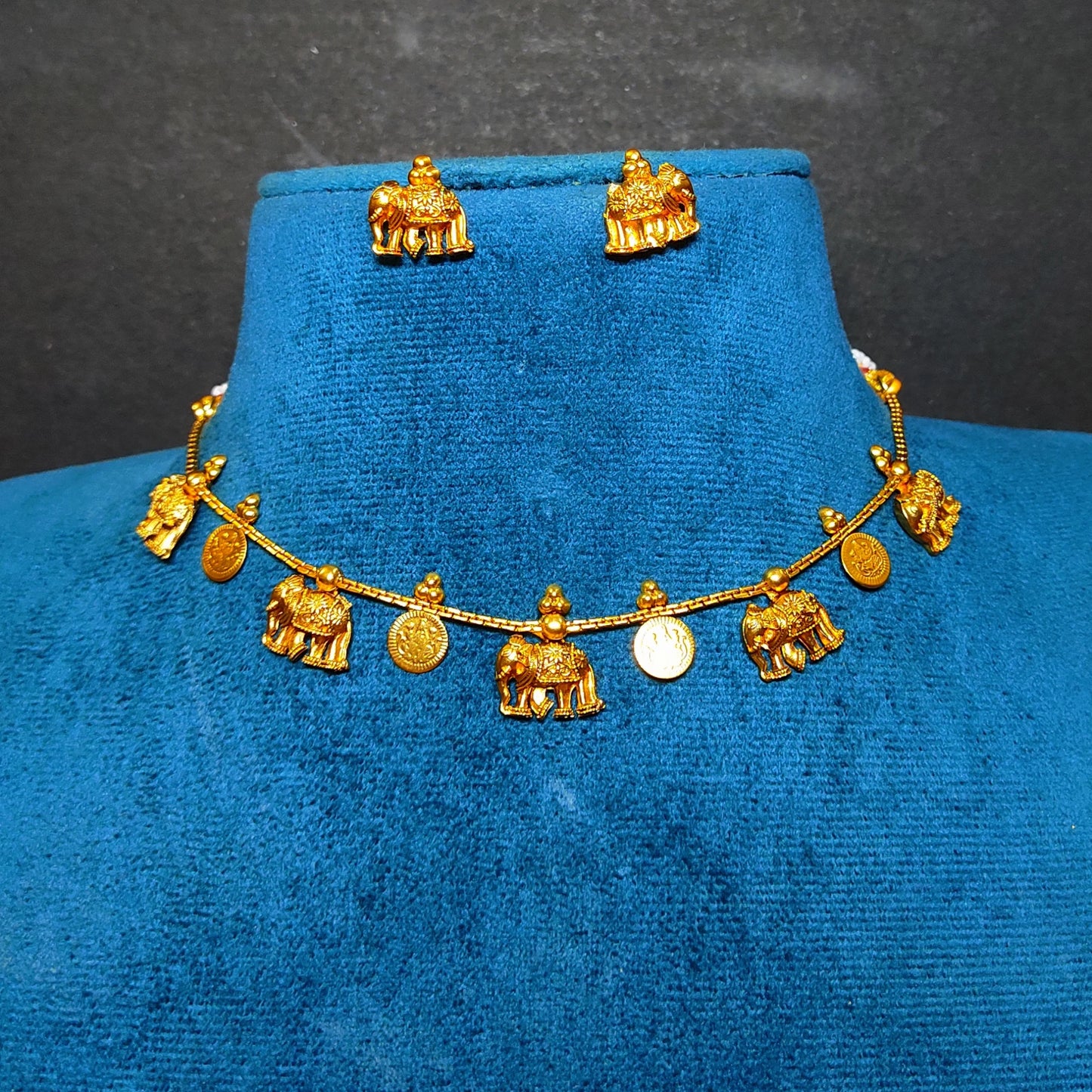 A gold-toned jewelry set displayed on a blue velvet bust, featuring a necklace with small elephant and coin-shaped charms and matching stud earrings.