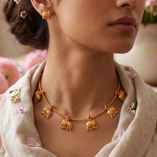 A close-up lifestyle shot of a woman wearing a gold toned elephant necklace and matching gold toned elephant stud earring, styled with a beige linen shawl and soft pink flowers.