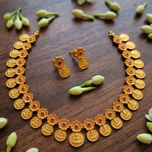 Antique Gold finish coin pendant necklace and matching stud earrings arranged on a dark wooden surface, decorated with small green flower buds. The jewelry features circular gold toned medallion pendants with red stone accents.