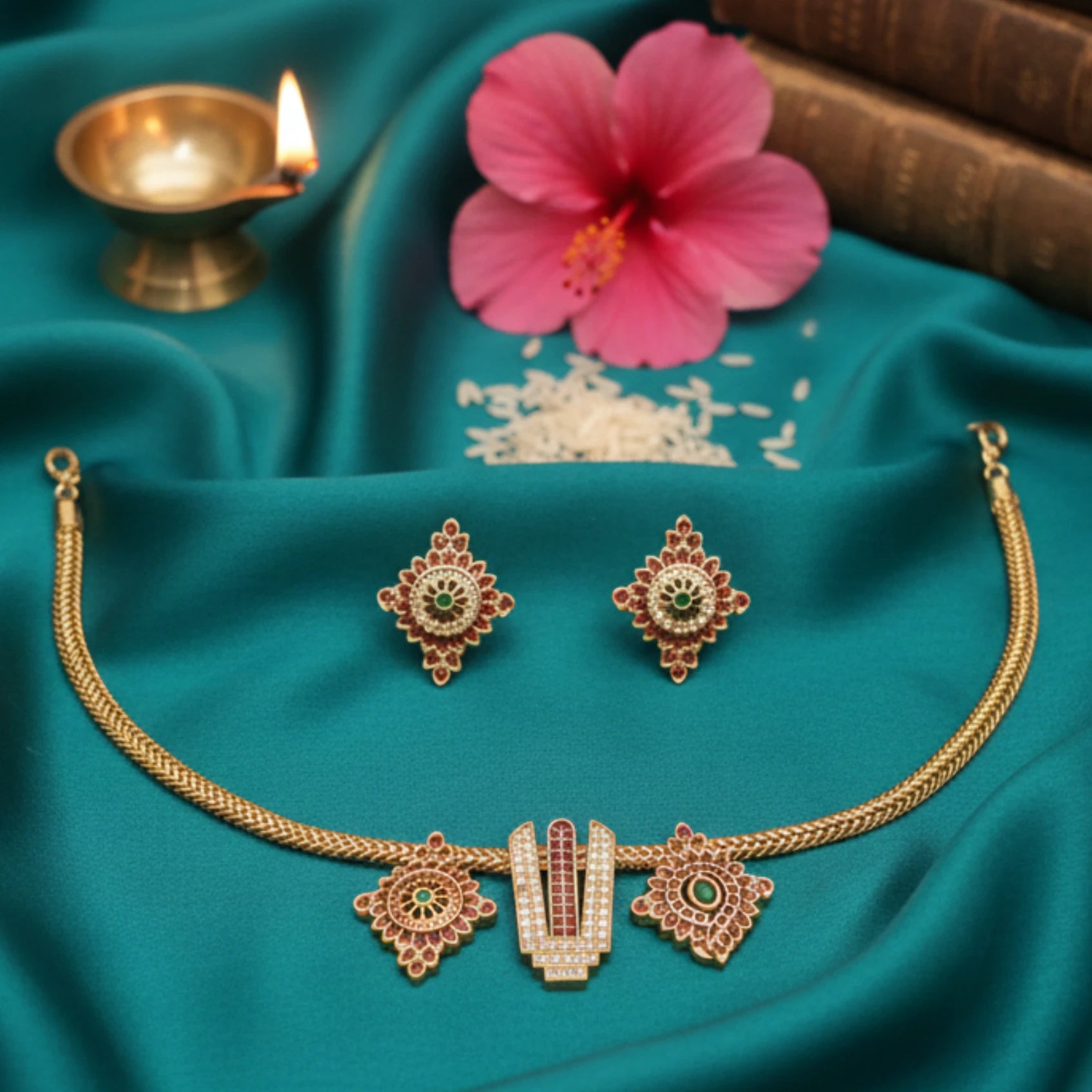 Gold toned necklace and earrings set on a teal fabric background with a pink flower and lit lamp.