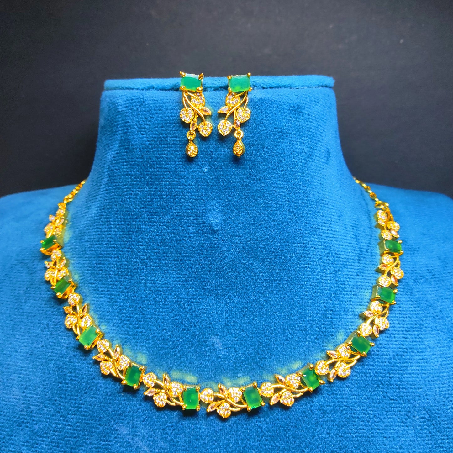 Gold plated necklace and earrings with green stones on a blue mannequin against a black background