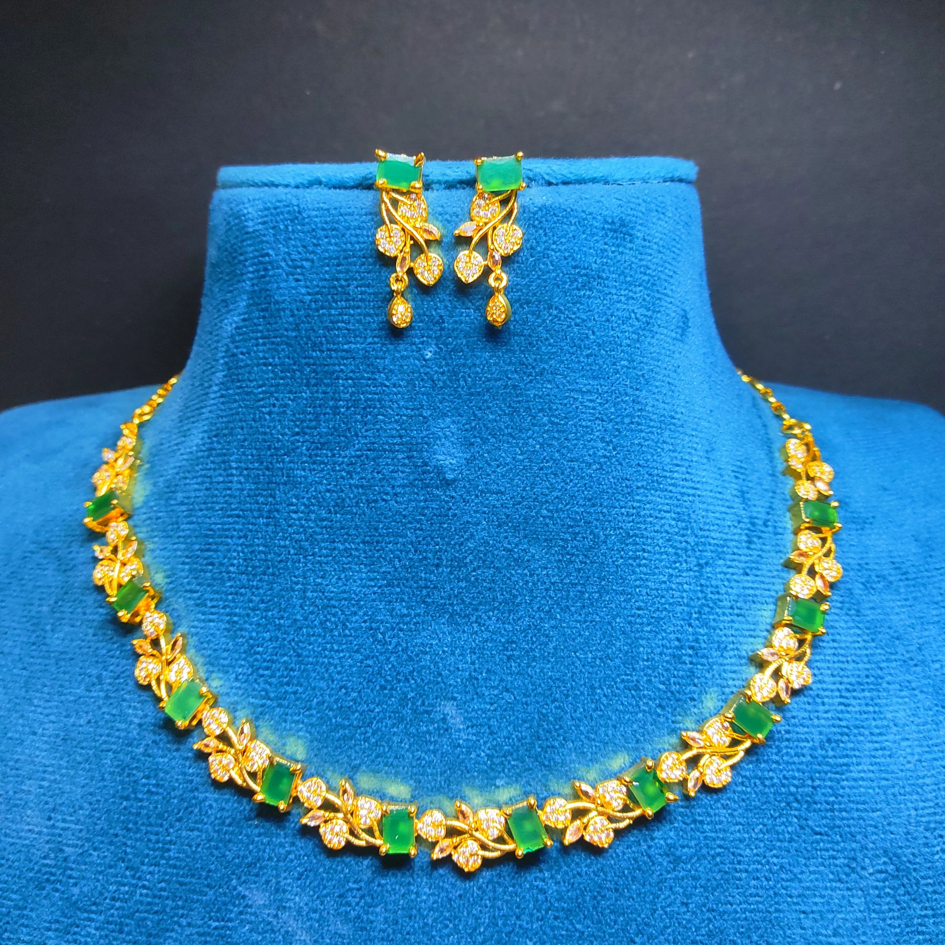 Gold plated necklace and earrings with green stones on a blue mannequin against a black background