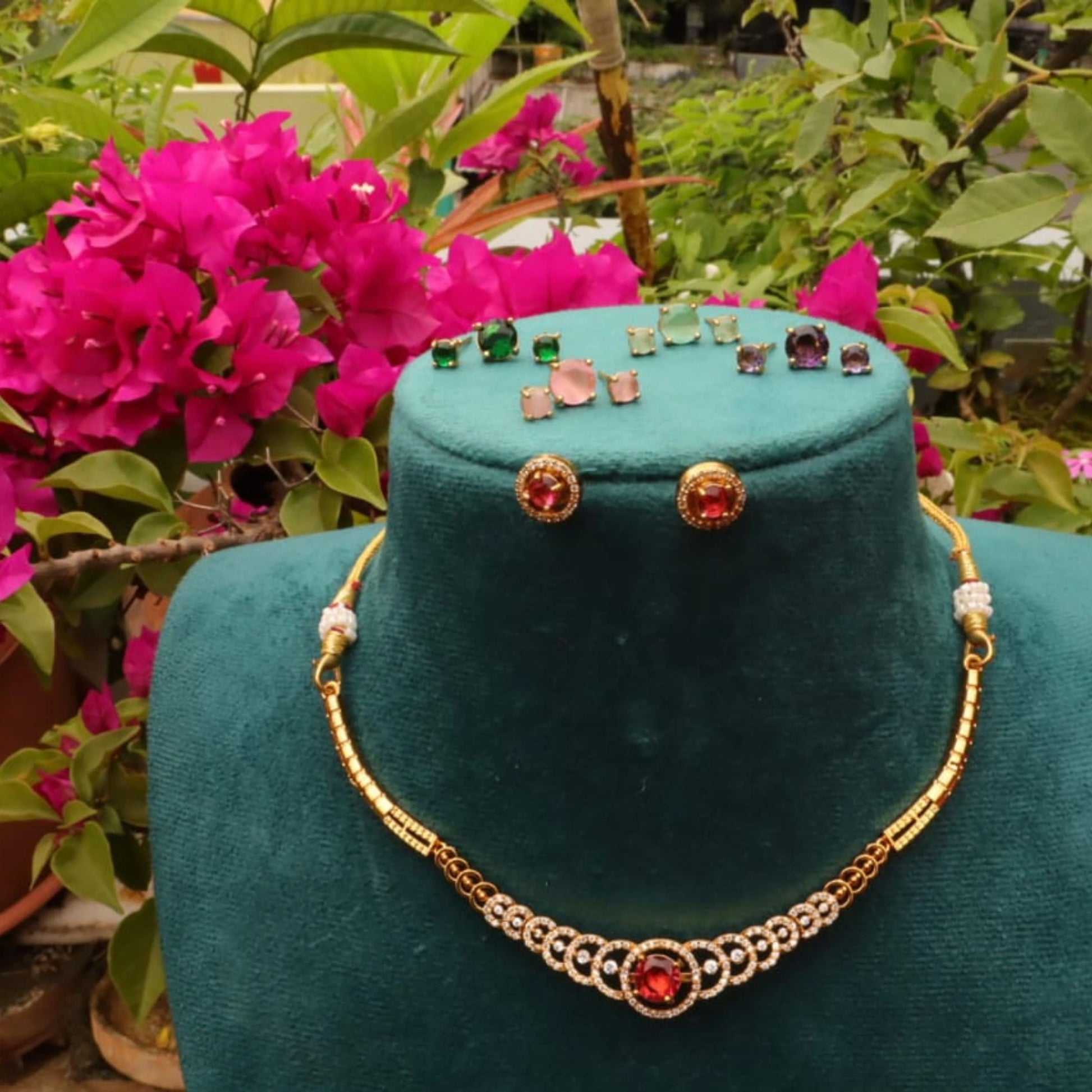 A gold-plated curved necklace with a red center stone and white accents, surrounded by five pairs of stud earrings in red, green, pink, blue, and teal in green mannequin