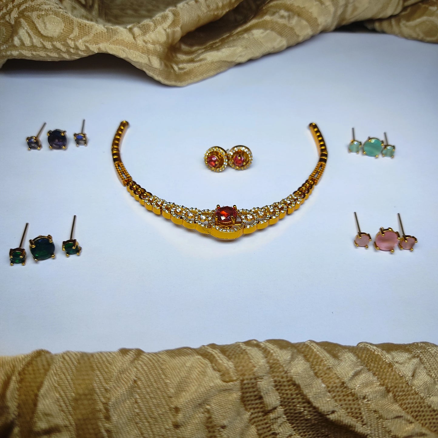 A gold-toned curved necklace with a red center stone and white accents, surrounded by five pairs of stud earrings in red, green, pink, blue, and teal