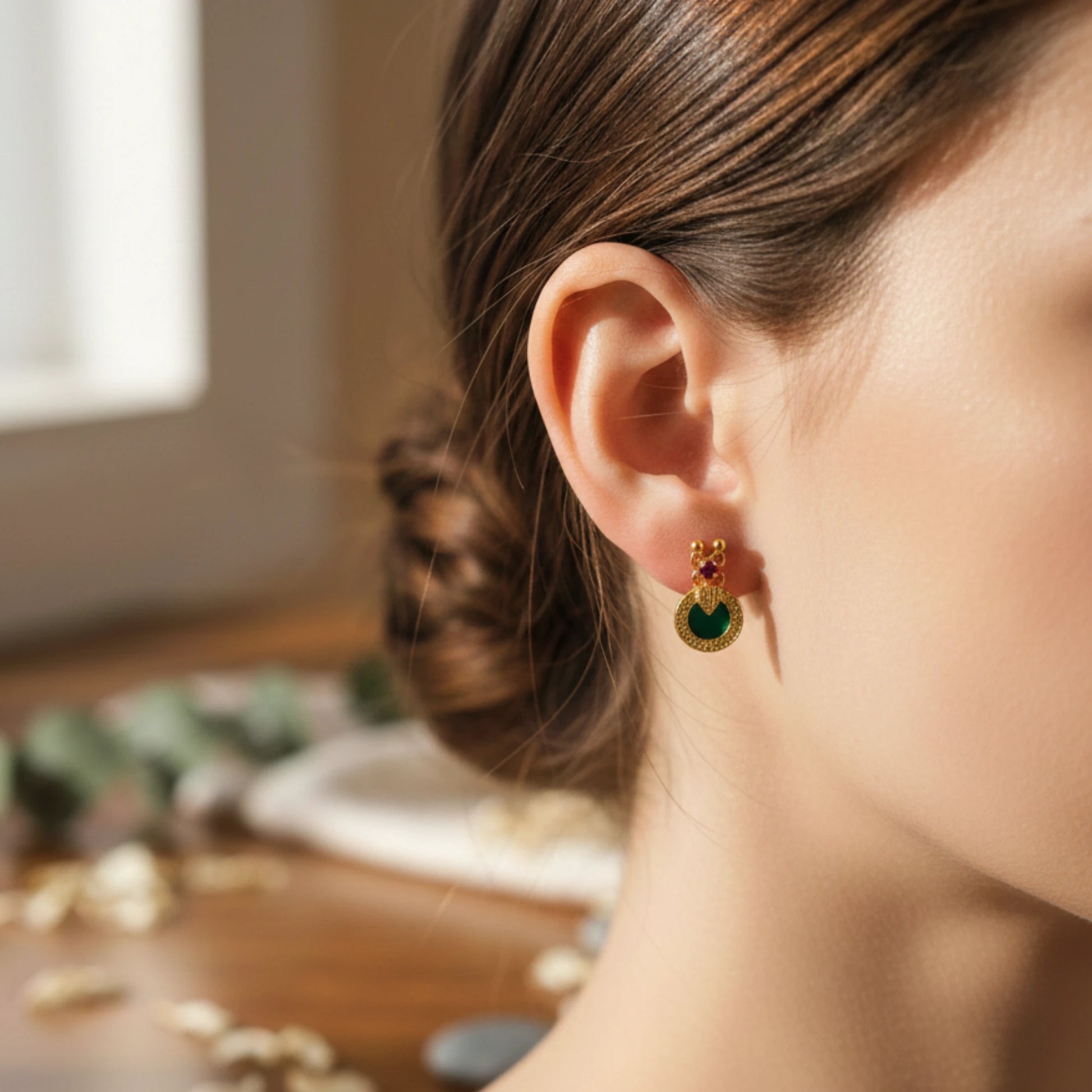 Close-up of a woman wearing a gold plated earring with a green palakka and ruby red gemstone, blurred background