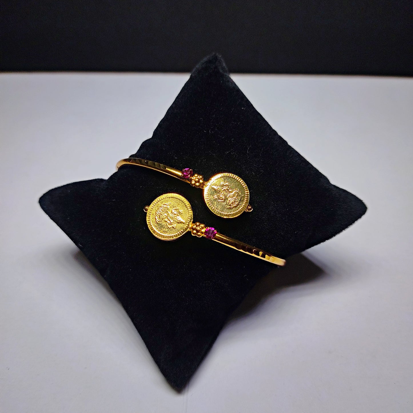 Gold plated bracelet with coin designs on a black pillow against a white background