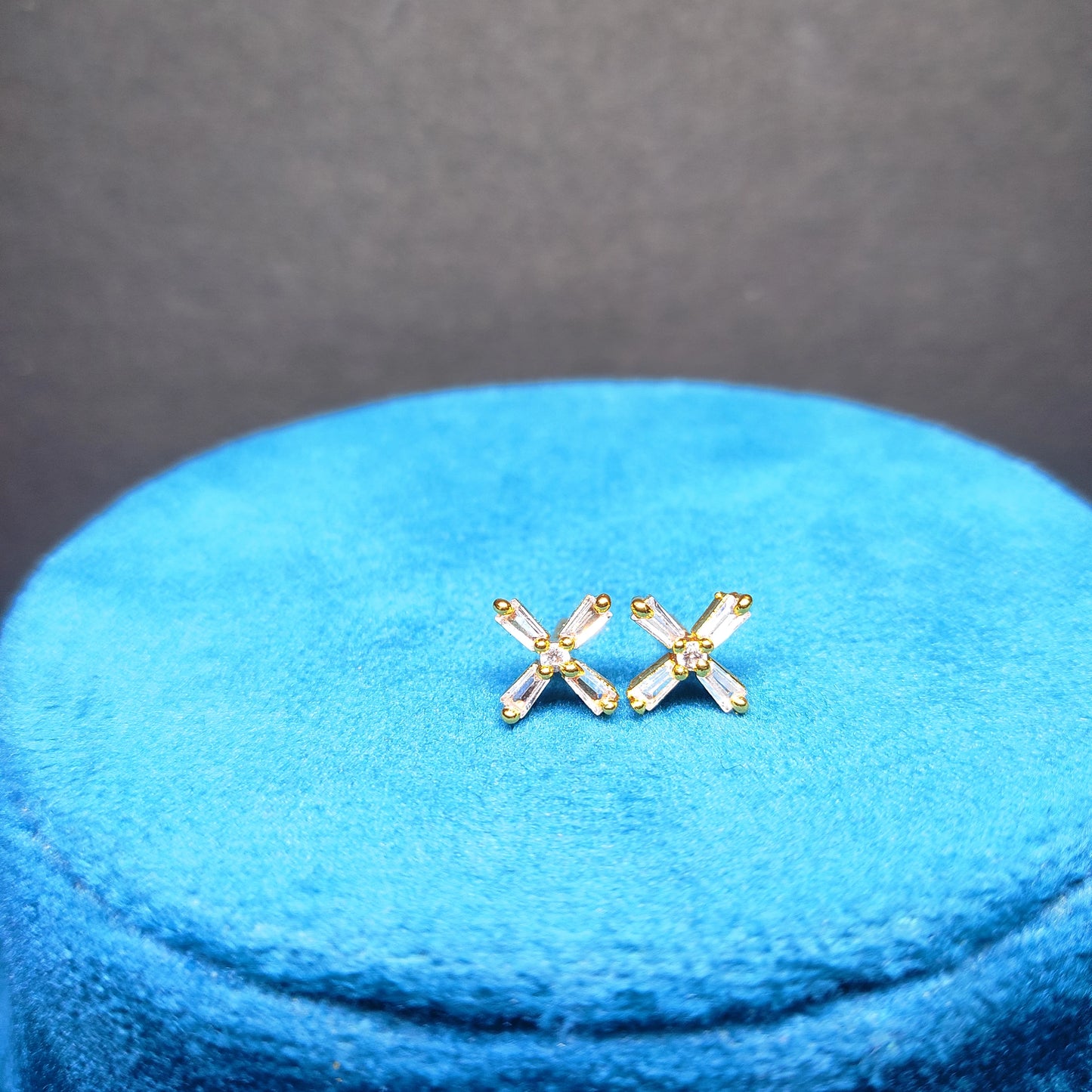 Gold plated 'X' shaped earrings on a blue cushion with a gray background
