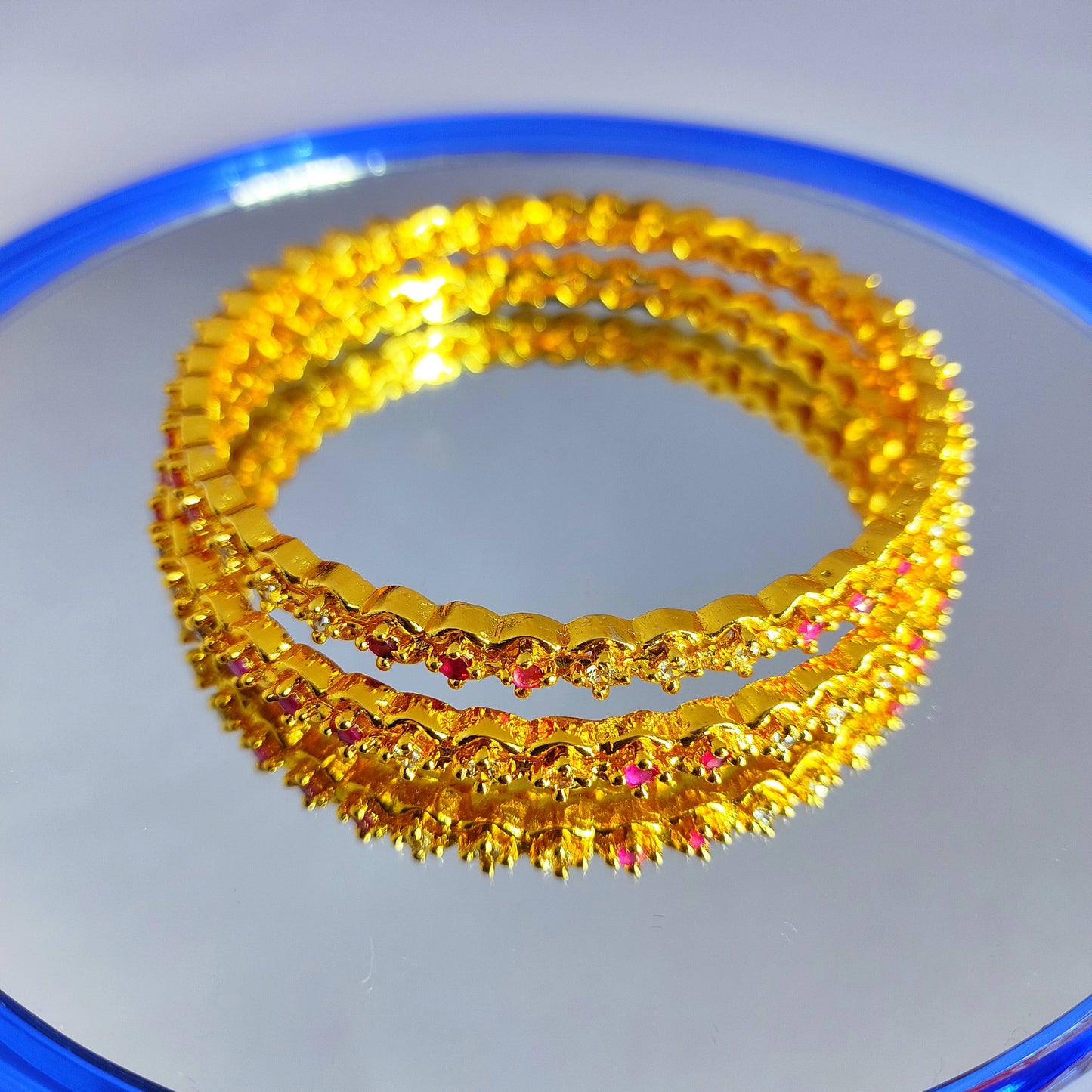 Two gold plated bangles with pink and white stones on a reflective surface