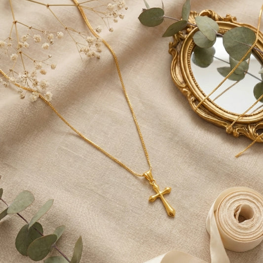 Gold plated neckchain with a cross pendant on a beige fabric background with greenery.