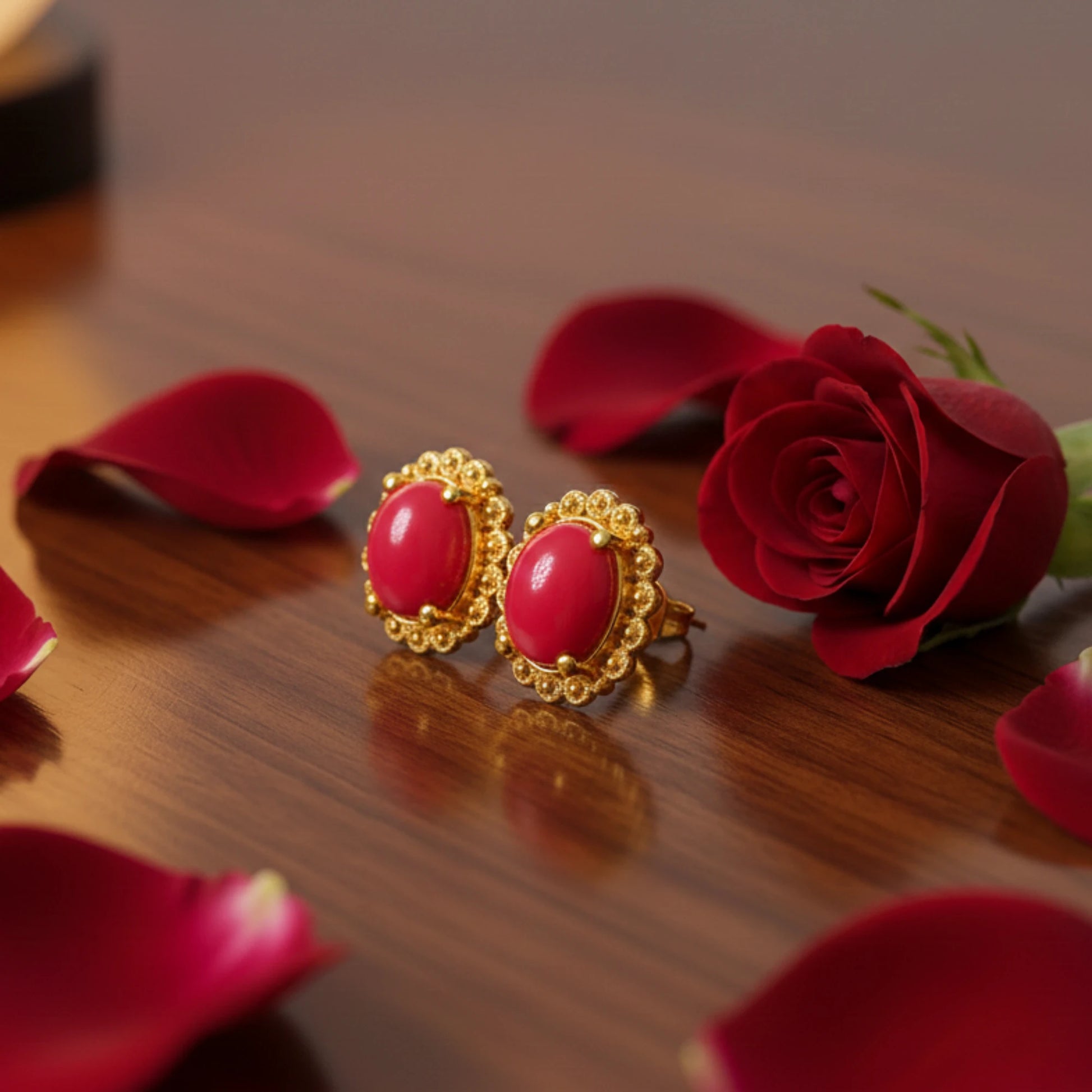 Gold plated earrings with oval red stones on a wooden surface with red roses and petals.