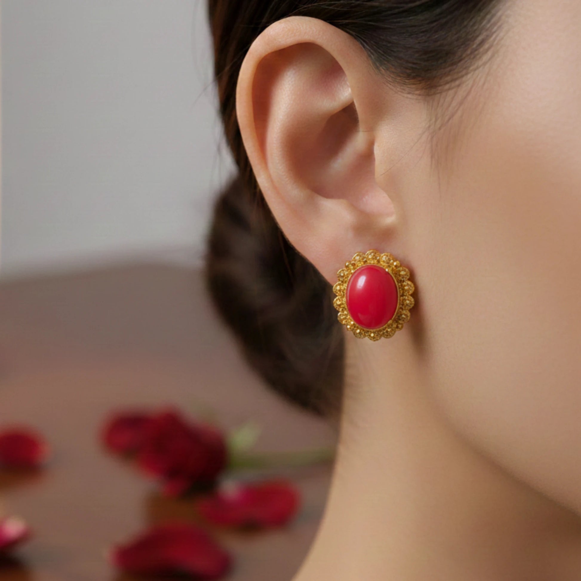Close-up of an ear wearing a oval red center earring with gold plating, blurred background with red flowers.