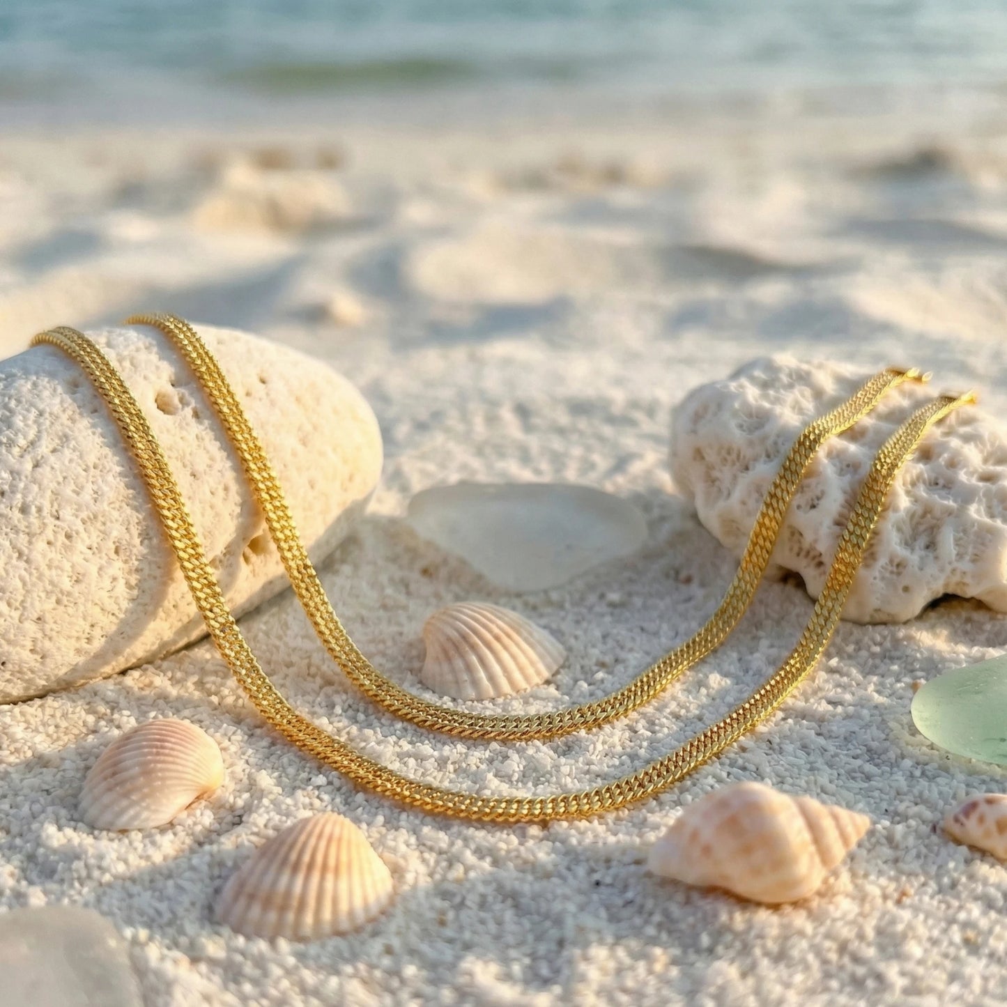 Gold plated anklets on a sandy beach with shells and rocks.