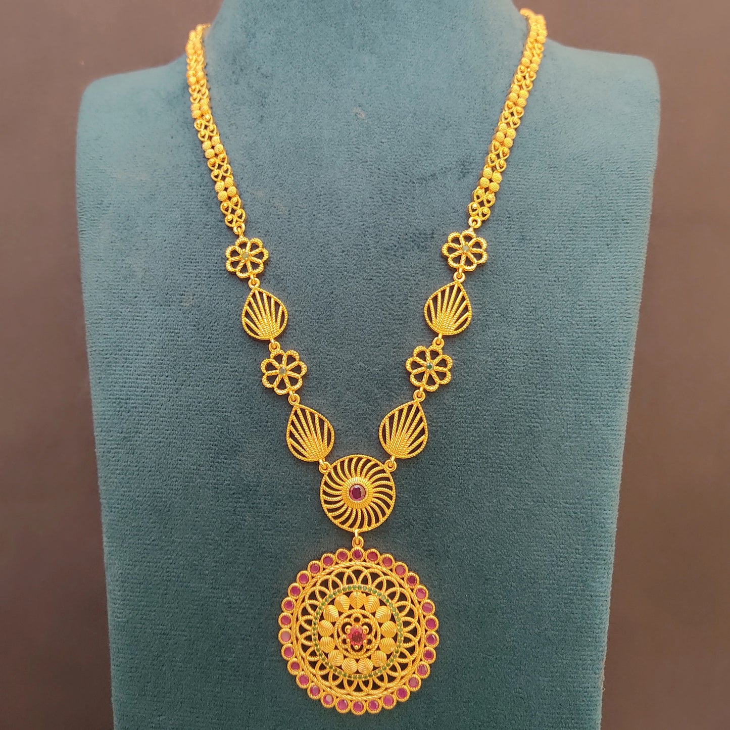Gold plated long ethnic necklace with intricate design and a floral mandala pendant on a green velvet stand against a brown background