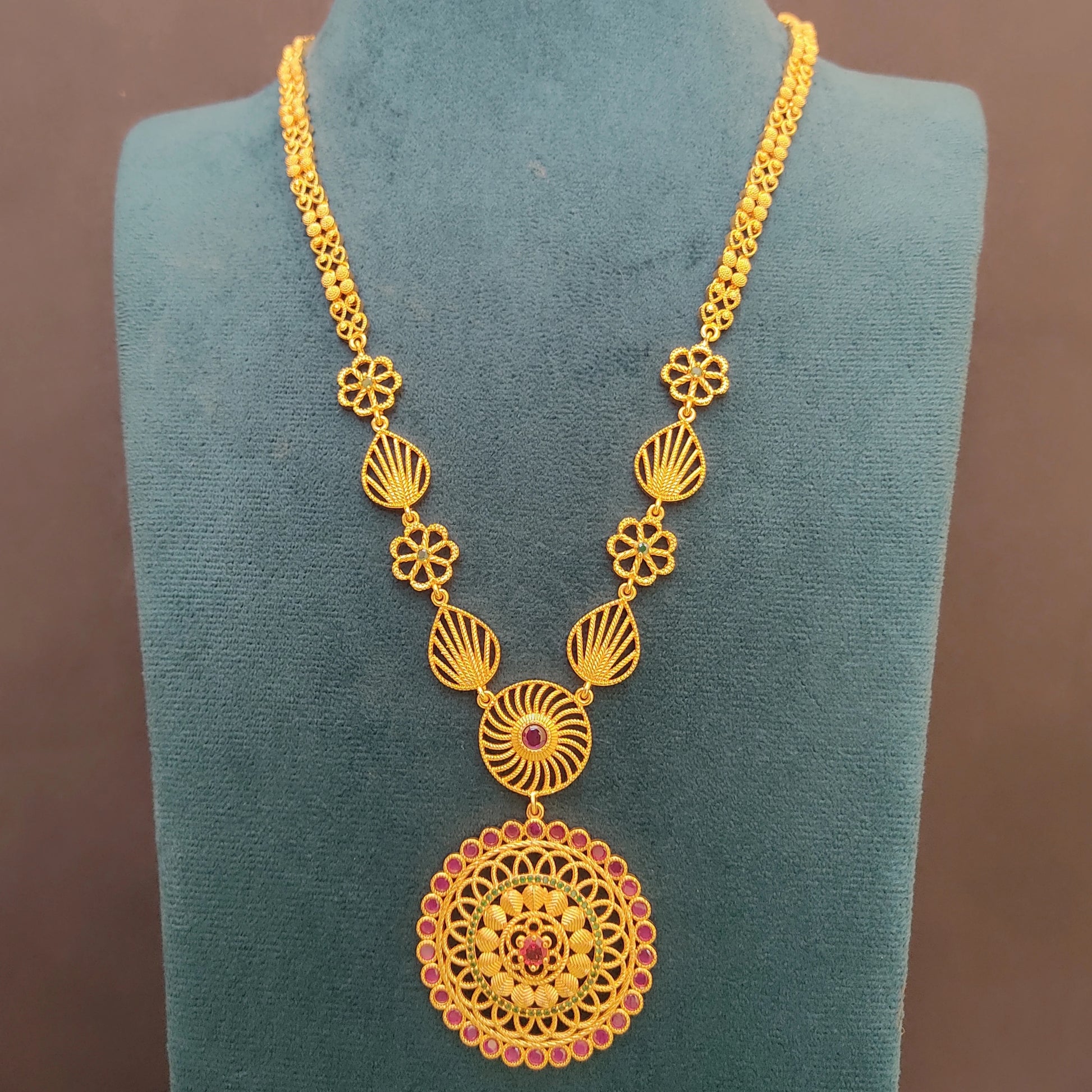 Gold plated long ethnic necklace with intricate design and a floral mandala pendant on a green velvet stand against a brown background