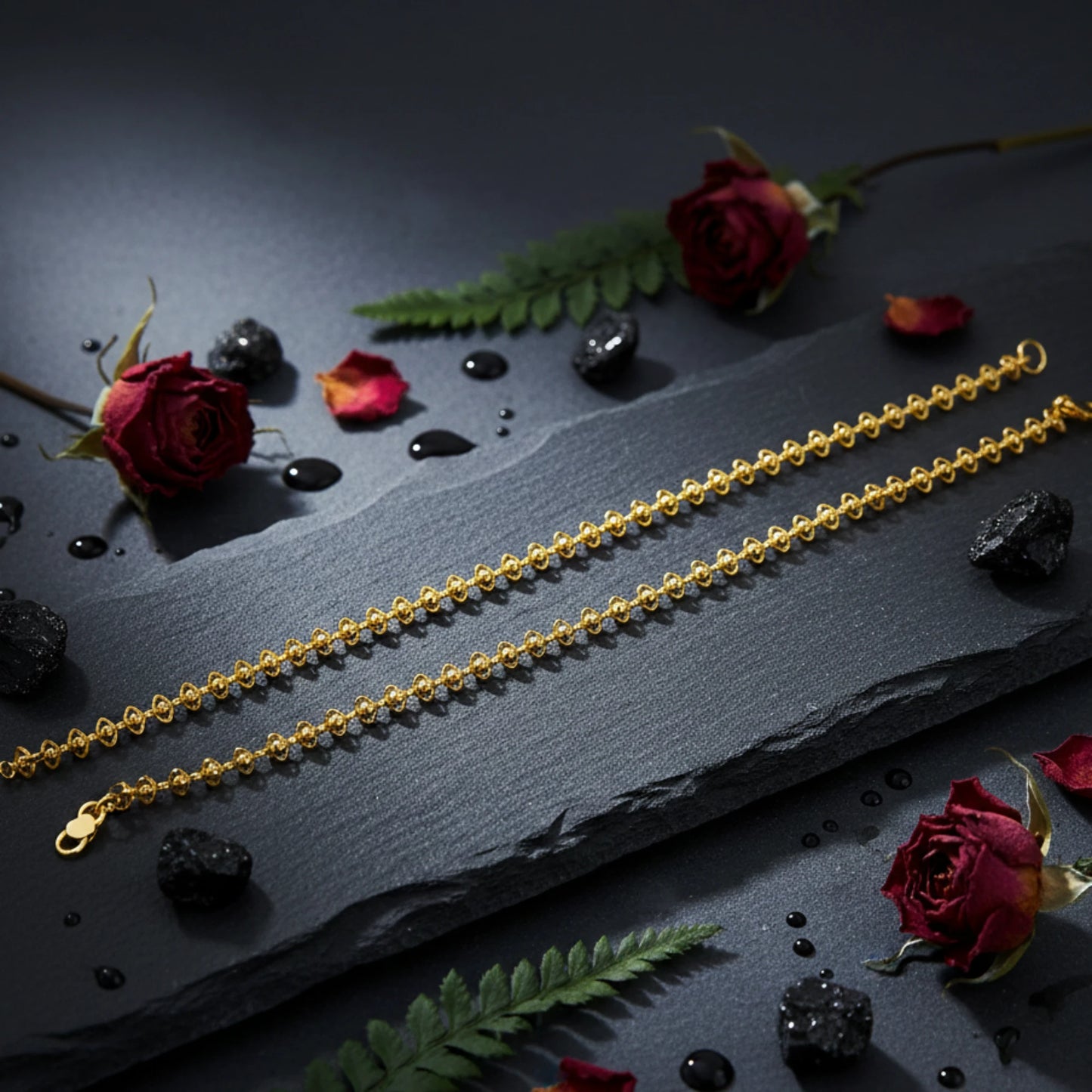Two gold plated delicate lace design anklets on a dark surface with red roses and green leaves.