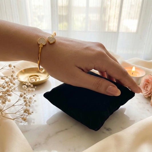 Hand with gold plated bracelet on a black pouch on a marble surface with flowers and candles.