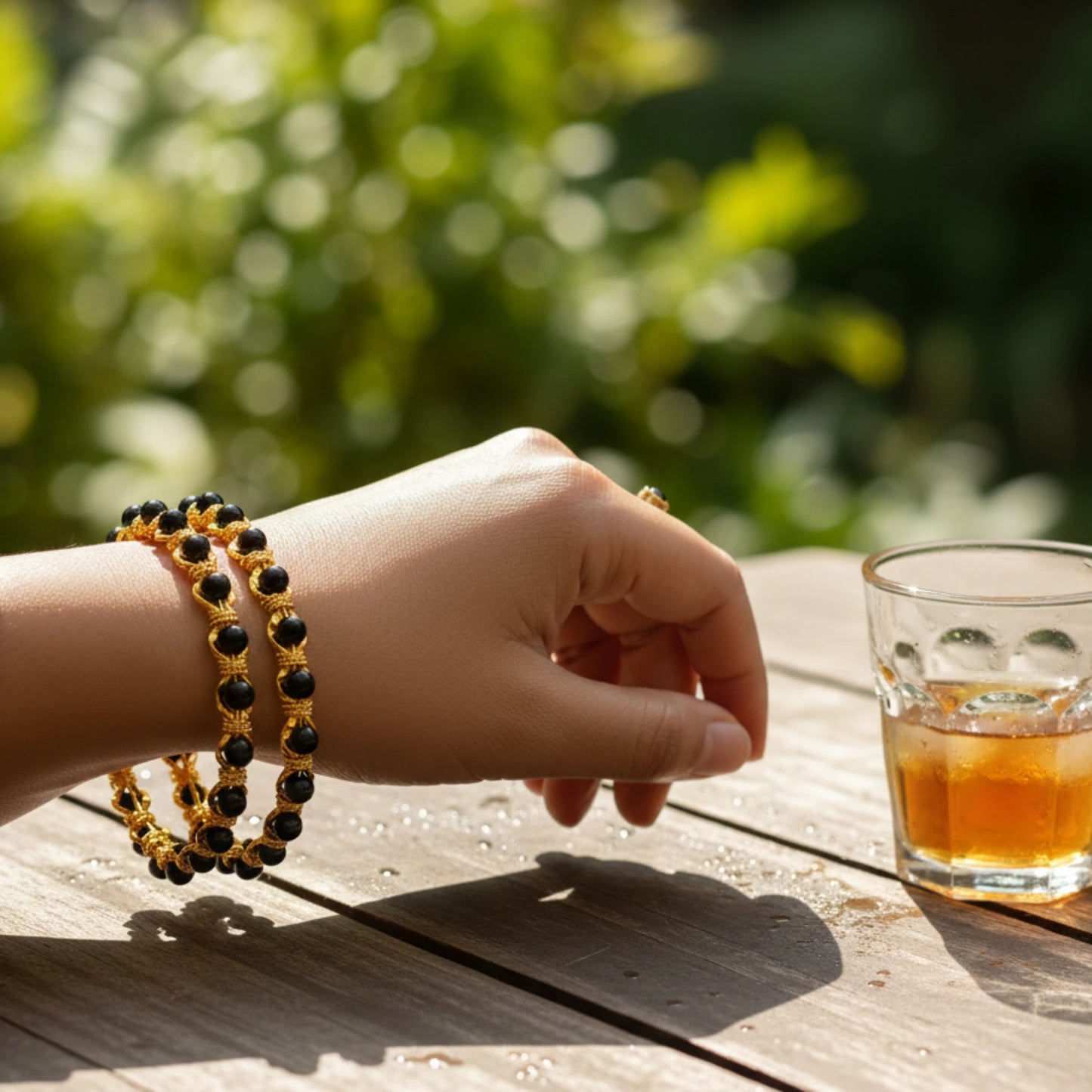Hand with gold plated bangles in karimani or black beaded on a wooden table with a glass of Tea.