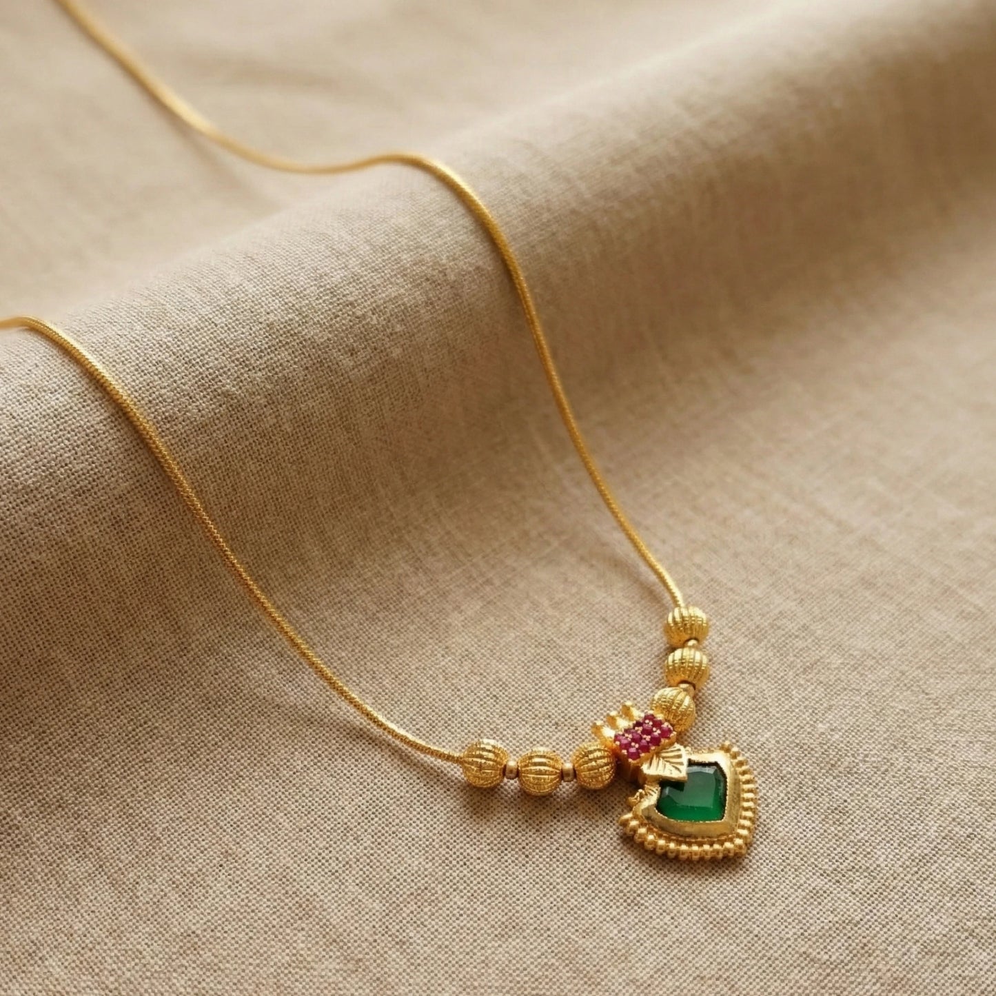 Gold plated necklace with a heart-shaped palakka pendant featuring green and red stones on a beige fabric background