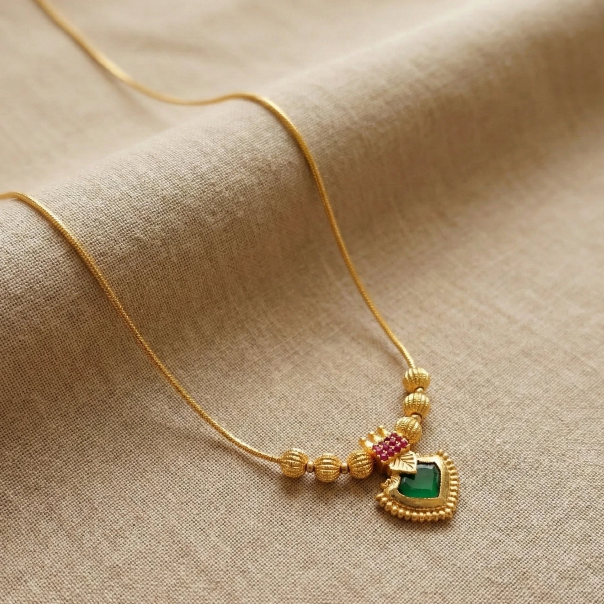 Gold plated necklace with a heart-shaped palakka pendant featuring green and red stones on a beige fabric background