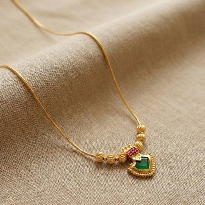 Gold plated necklace with a heart-shaped palakka pendant featuring green and red stones on a beige fabric background