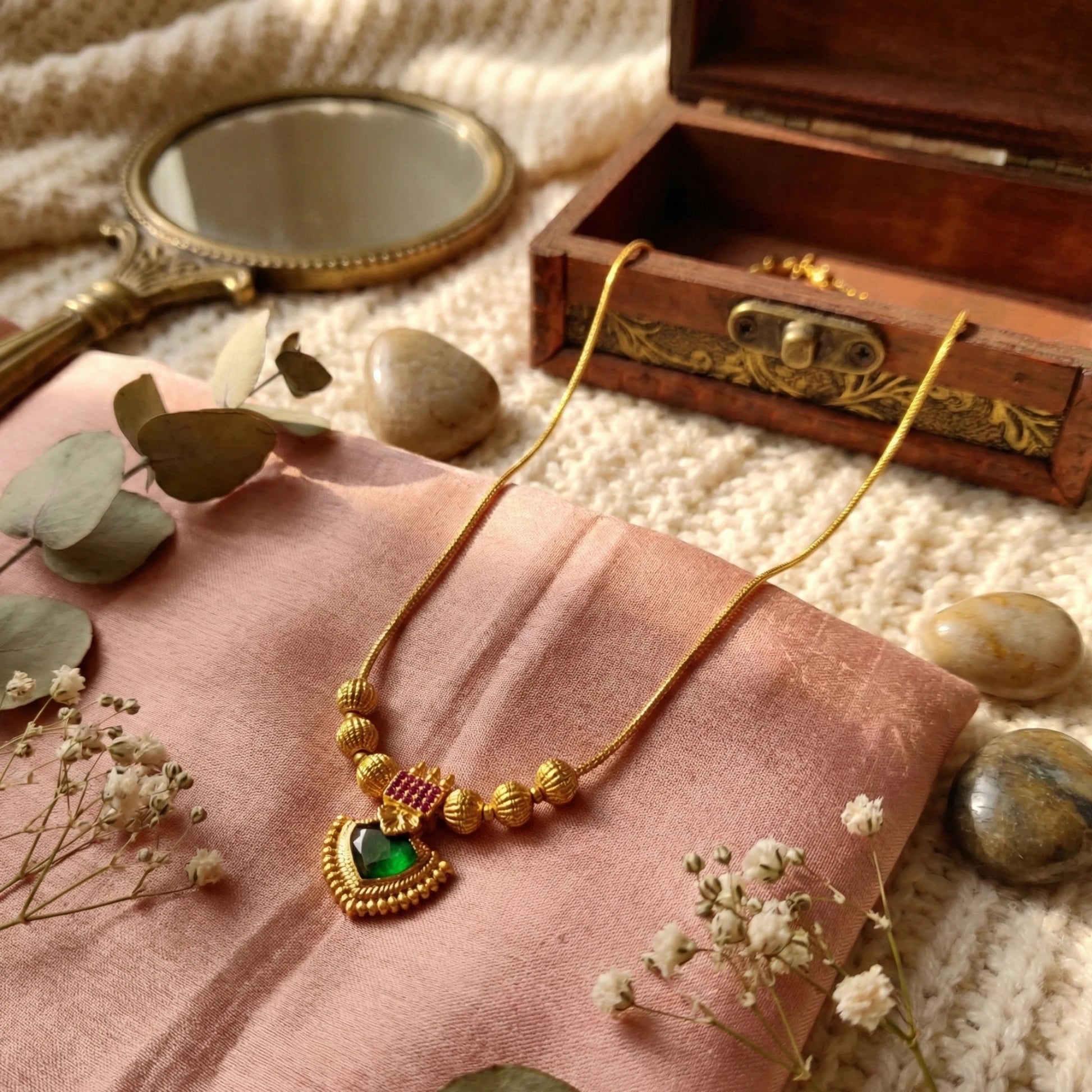 Gold plated palakka necklace with green palakka on pink fabric, surrounded by stones and a mirror.